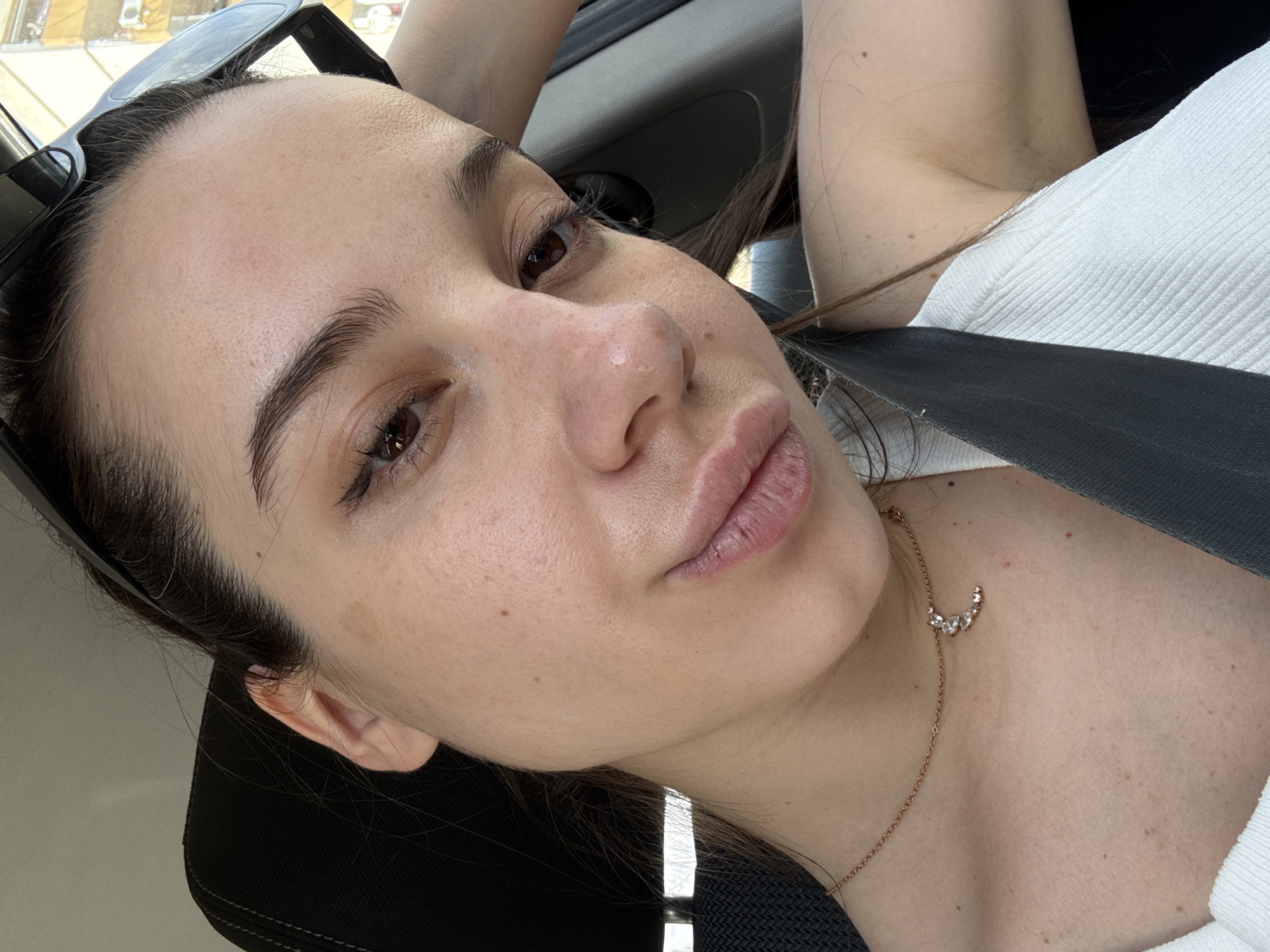 A close-up, angled selfie depicts a young woman with fair skin, brown hair, and brown eyes. She is wearing a white, textured top and a gold necklace with a crescent-shaped pendant of small, clear stones. A black car seatbelt crosses her chest. Sunglasses are positioned on her head. The background shows the interior of a car and a glimpse of an exterior urban environment, possibly in Sofia, Bulgaria. The woman is looking directly into the camera.