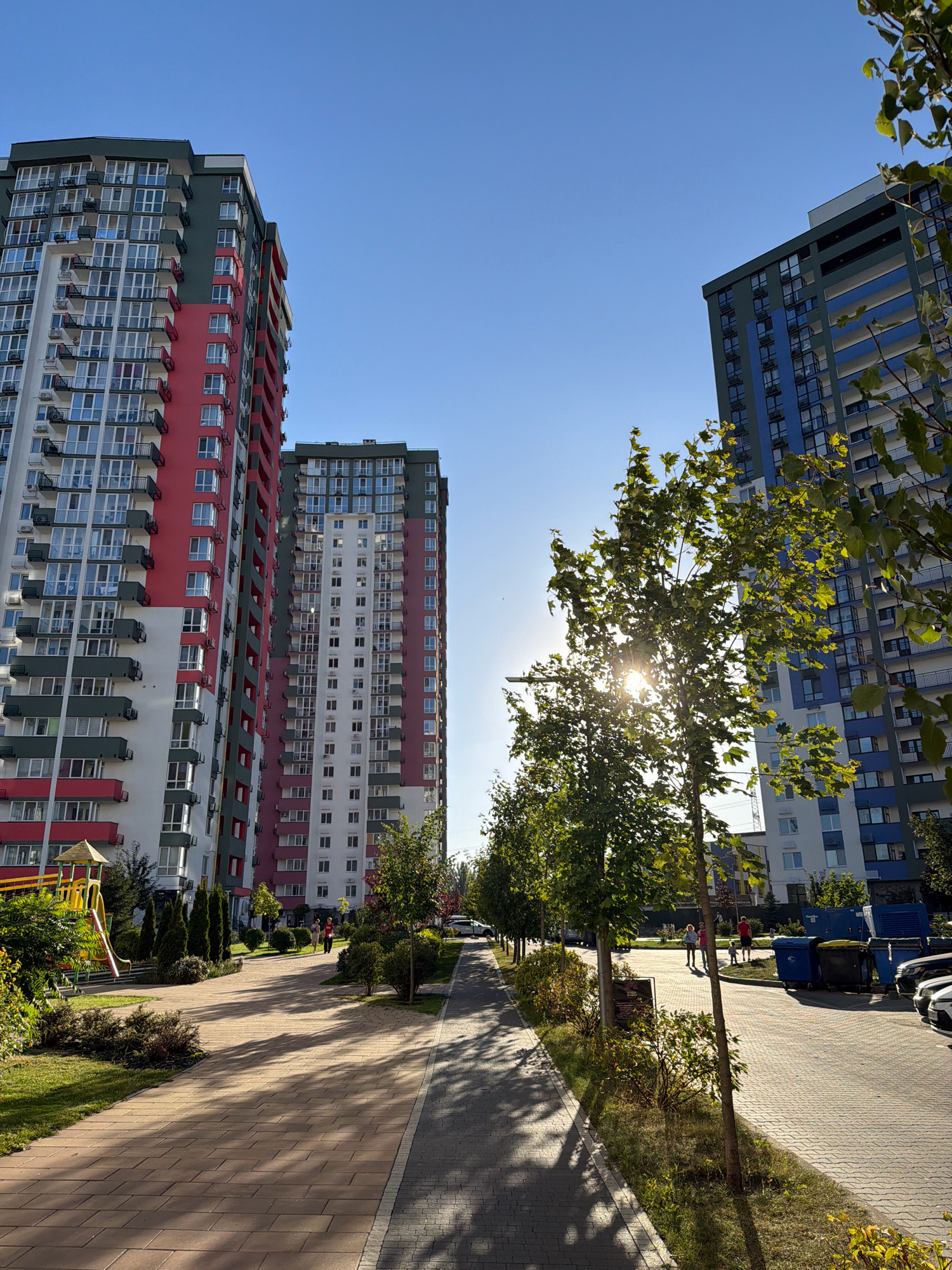 The image depicts a sunny day in a modern residential complex in Kyiv, Ukraine. In the foreground, a paved walkway with a pattern of light brown and gray interlocking stones runs alongside a grassy area with neatly trimmed bushes and small trees. The walkway is partially covered in dappled shadows cast by trees lining the path.
Dominating the background are several tall, contemporary apartment buildings. The building on the far left is predominantly white with prominent sections of bright red and dark grey. It features numerous balconies with dark railings and a playground with a yellow slide and a red roofed structure on its lower left side. To its right, another apartment building shares a similar architectural style, featuring white and red vertical sections. Further to the right, a third building is characterized by its blue and white facade, also with multiple balconies. The sky is a clear, bright blue, indicating good weather conditions.
On the walkway, two adults and a child are walking away from the camera towards the right side of the frame. On the far right of the image, a row of large blue industrial bins and several parked cars are visible, suggesting a communal waste disposal area and parking. The presence of modern buildings, well-maintained landscaping, and a playground indicates a contemporary urban residential environment.