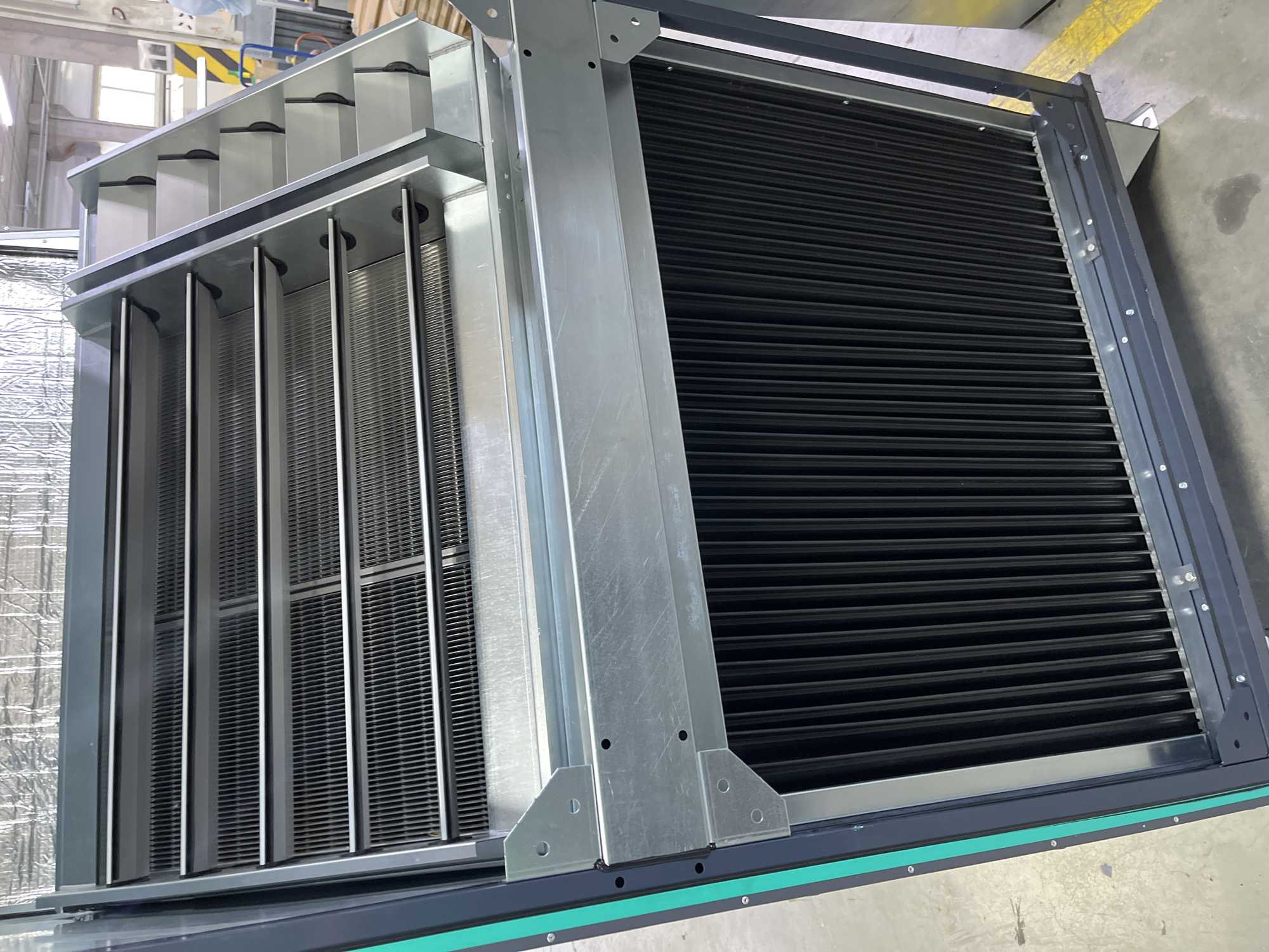 An industrial air handling unit is shown in a warehouse setting. The unit is a large, rectangular metal structure with several distinct sections. The top section appears to be a filter or heat exchanger, with multiple rows of thin, parallel slats. Below this is another section with a dark, ribbed surface, likely another filter or a collection of finned tubes. The overall structure is dark gray with some bright green trim along the left edge. The unit is standing on a concrete floor with yellow painted lines, typical of industrial or warehouse environments. In the background, there are glimpses of other industrial equipment, including metal shelving, a blue wheeled cart, and possibly some wooden pallets. The lighting suggests an indoor, well-lit space. The image does not depict any people or specific actions, focusing solely on the industrial equipment. There is no information in the image to determine the location in Kyiv, Ukraine.