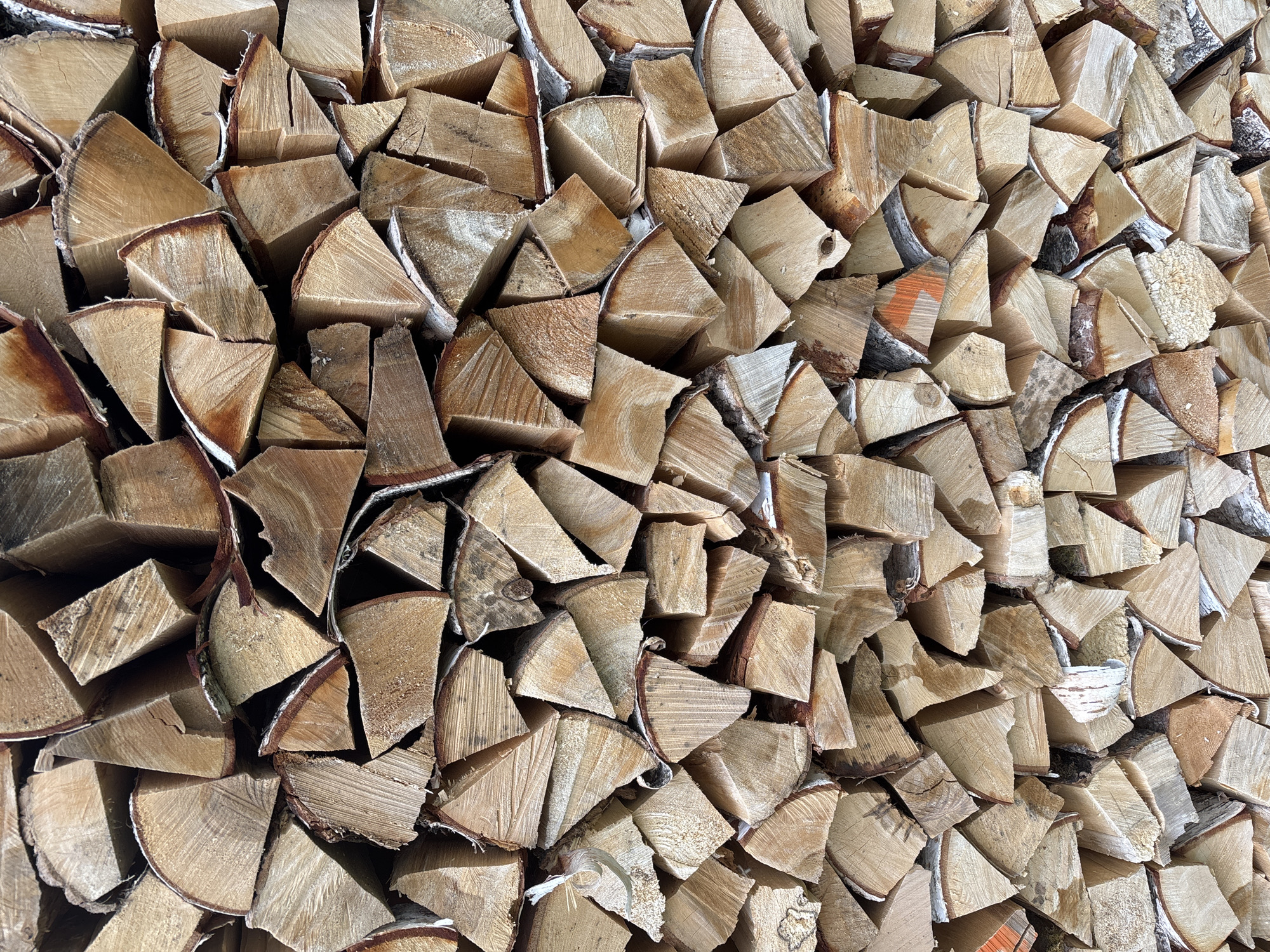The image is a close-up, high-angle, full-frame shot of a neatly stacked pile of firewood. The wood appears to be birch, with light-colored bark peeling in places and the exposed wood grain showing variations in light brown and cream tones. The logs are cut into uniform lengths and are stacked in an interlocking pattern, creating a dense wall of wood. Some pieces show visible bark, while others are cleanly cut, revealing the wood's texture. The lighting suggests an outdoor environment, with shadows and highlights emphasizing the contours and textures of the wood. There are no people or animals visible in the image. The location in Kurgan, Russia is not determinable from the visual information provided in the image.