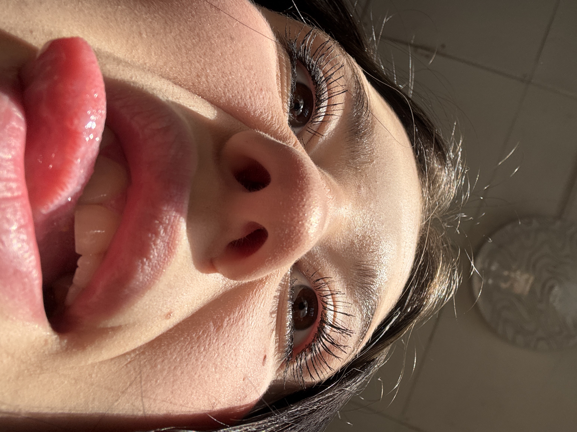 A close-up, low-angle shot focuses on a woman's face with her tongue extended. The woman has fair skin, brown eyes, and dark hair. She wears dark mascara, emphasizing her eyelashes. The woman's nose is centered in the frame, and her mouth is slightly open, revealing her front teeth. The tongue is prominent, filling the left side of the image. The background is blurred and indistinct. The location is not discernible from the image.