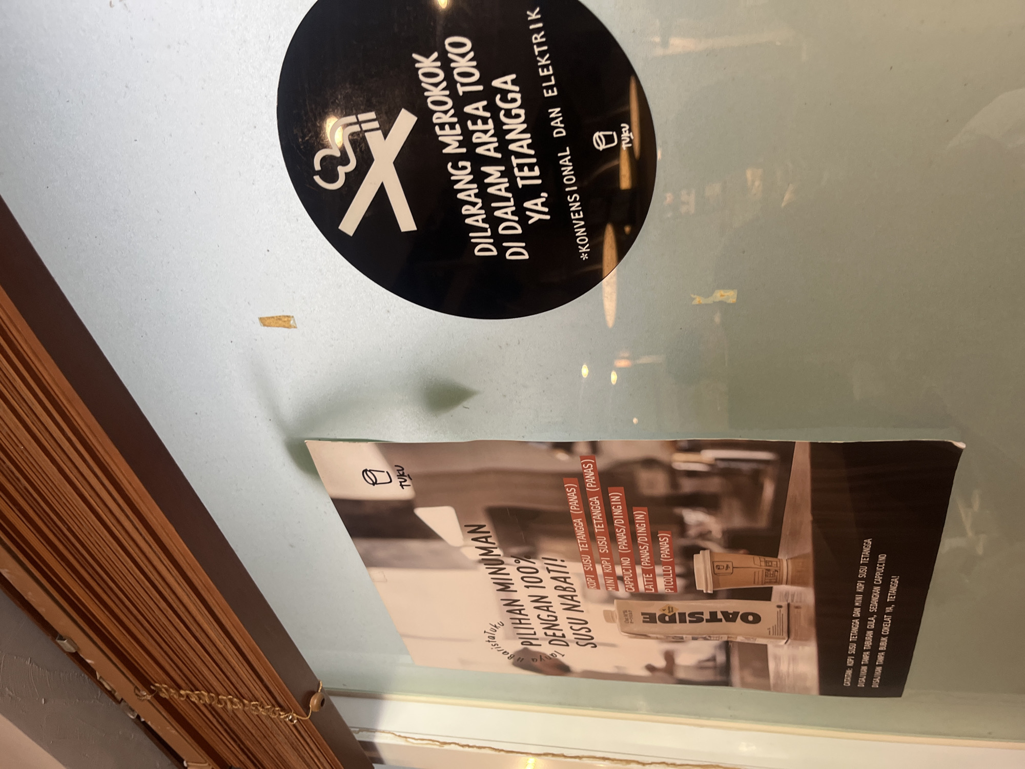 A poster promoting oat milk beverages is affixed to a light blue wall next to a dark circular sticker with a no-smoking symbol and text in Indonesian. The poster, partially obscured by a wooden blind, displays the title "PILIHAN MINUMAN DENGAN 100% SUSU NABATI!" which translates to "CHOICE OF DRINKS WITH 100% PLANT MILK!". Below this are listed various coffee drinks with oat milk options, such as "KOPI SUSU TETANGGA (PANAS)" (Neighbor's Milk Coffee (Hot)), "MINI KOPI SUSU TETANGGA (PANAS)" (Mini Neighbor's Milk Coffee (Hot)), "CAPPUCCINO (PANAS/DINGIN)" (Cappuccino (Hot/Cold)), "LATTE (PANAS/DINGIN)" (Latte (Hot/Cold)), and "PICCOLLO (PANAS)" (Piccolo (Hot)). A smaller text at the bottom of the poster provides notes on specific drinks, including "CATATAN: KOPI SUSU TETANGGA DAN MINI KOPI SUSU TETANGGA DISAJIKAN TANPA TABURAN GULA, SEDANGKAN CAPPUCCINO DISAJIKAN TANPA BUBUK COKELAT YA, TETANGGA!" (Note: Neighbor's Milk Coffee and Mini Neighbor's Milk Coffee are served without sugar sprinkles, while Cappuccino is served without chocolate powder, neighbor!). The dark circular sticker features a white no-smoking symbol with a cigarette and smoke crossed out. Below this, the text reads "DILARANG MEROKOK DI DALAM AREA TOKO YA, TETANGGA" (SMOKING IS PROHIBITED IN THE STORE AREA, NEIGHBOR) and "*KONVENSIONAL DAN ELEKTRIK" (*CONVENTIONAL AND ELECTRIC). Both the poster and the sticker include the logo of a simple line drawing of a cup with the word "TUKU" below it. The environment appears to be indoors, likely a cafe or food establishment, with reflections visible on the blue wall. The overall impression is a promotional and informational display within a commercial space.
