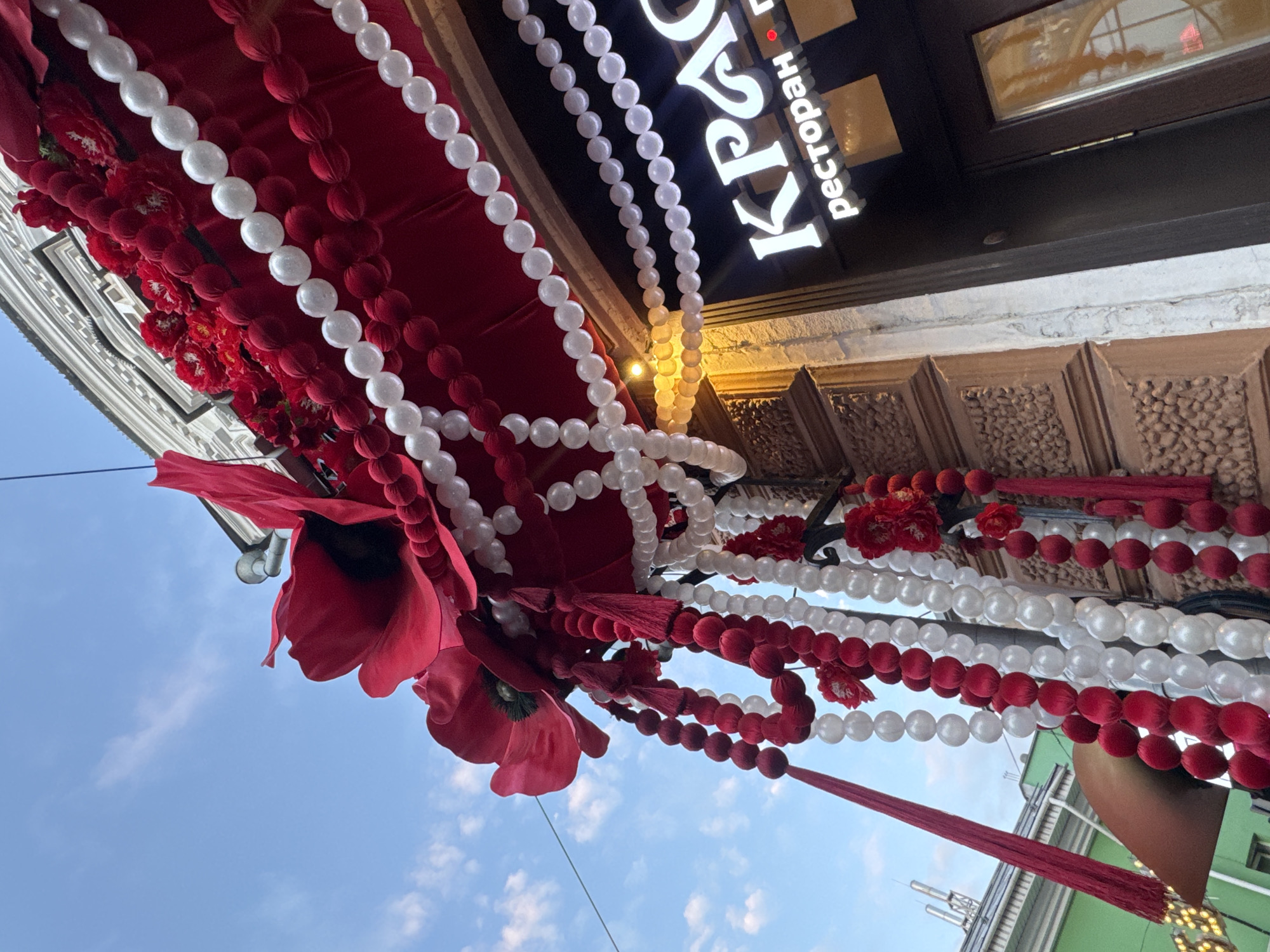The image shows a festive outdoor decoration in front of a restaurant in Nizhny Novgorod, Russia. The decoration features large red poppies, strings of red and white beads, and red tassels hanging from an awning. The awning itself is red, adorned with more red flowers and bead strings. Above the awning, a portion of a building with ornate architectural details is visible, along with a blue sky with scattered clouds. A sign for "КРАС" (likely part of a restaurant name) and "ресторан" (restaurant) is illuminated on the building's facade. The overall scene suggests a celebratory or decorative display, possibly for an event or season.