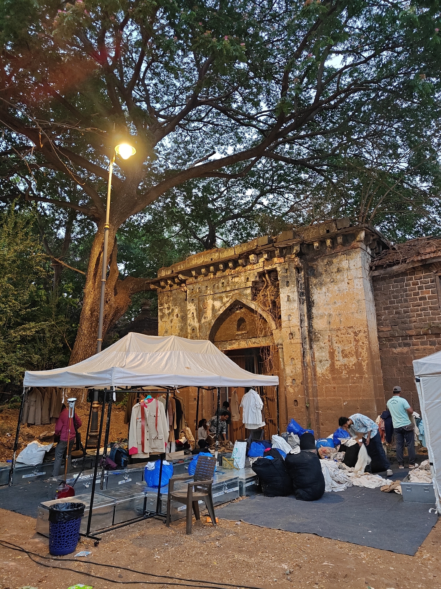 The image depicts an outdoor scene at dusk or early evening in Kolhapur, India, situated adjacent to an ancient, weathered stone building. This structure, possibly a historical gateway or fort wall, features an arched entrance with intricate stone carvings, corbels, and visible signs of disrepair, including erosion and roots growing from its facade. A very large, mature tree with an extensive canopy dominates the upper left portion of the frame, with its branches sprawling across the sky.
Beneath the tree and partially obscuring the historical building, a tall streetlamp with a bright yellow light source illuminates the immediate area. In the foreground, a temporary white canopy tent is erected, housing clothing racks displaying various garments, including jackets and shirts. Beneath this tent, multiple silver metal storage trunks are arranged on a dark ground mat. To the left of the tent, a blue perforated waste bin and a wooden chair are visible.
Several individuals are present, engaged in activities related to the handling of textiles. One person in a maroon shirt stands near the left side of the tent. Further right, a person in a light blue patterned shirt is seen bending over a pile of clothing on the ground. Another individual in a light blue t-shirt stands facing away from the viewer near a smaller, secondary white tent or canopy on the far right. Numerous bags, primarily blue and black, containing clothing or fabric are scattered on the ground mats, interspersed with piles of loose textiles. Black cables lie across the dirt ground in the foreground. The overall impression is that of a temporary setup, possibly for a market stall or a costume department.