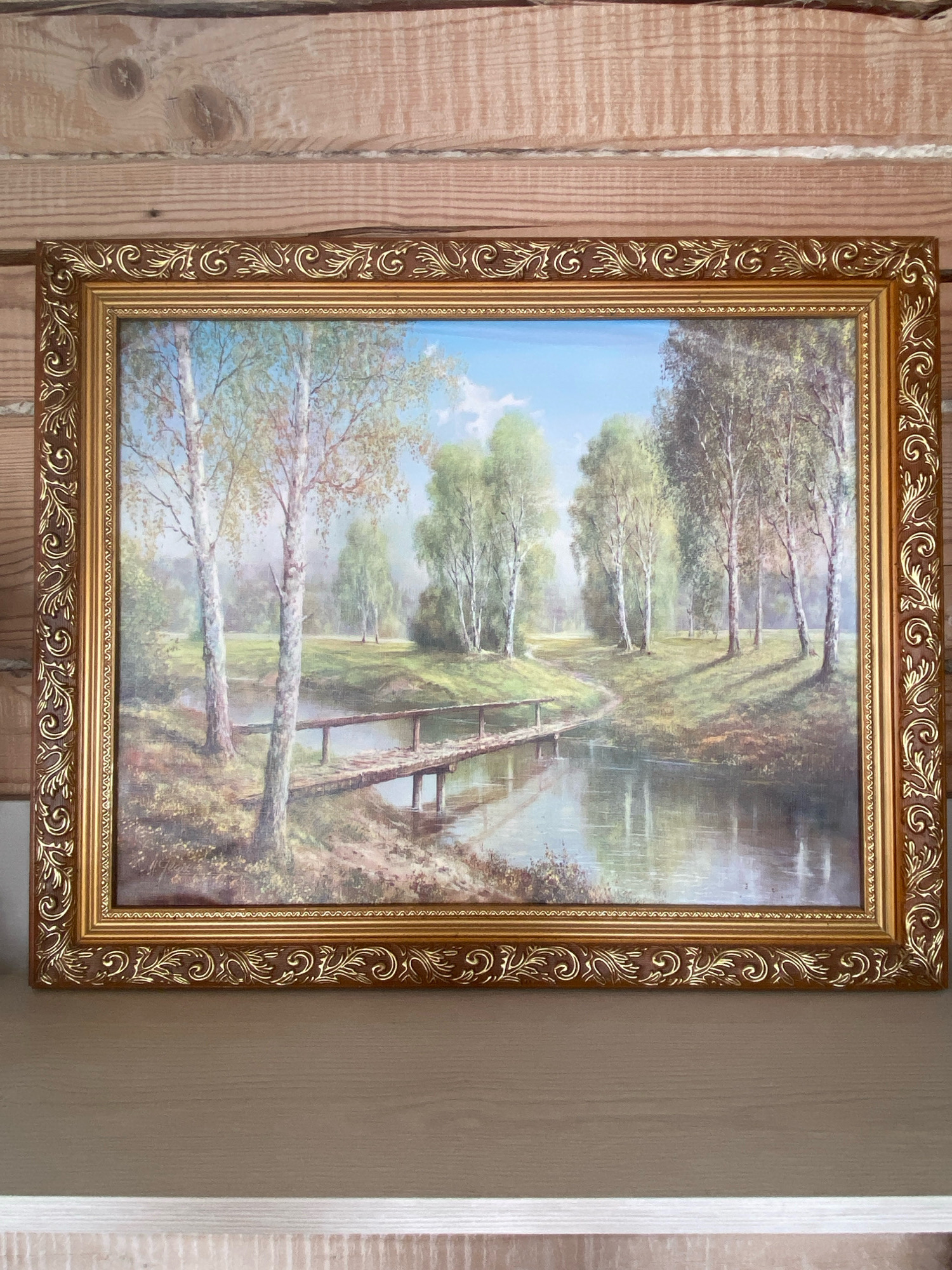 A framed painting of a serene landscape is displayed against a wooden paneled wall. The painting depicts a tranquil scene with a body of water reflecting the sky and surrounding trees. A rustic wooden bridge crosses the water, leading to a path that winds through a sun-dappled clearing. Several birch trees with their characteristic white bark are prominent in the foreground and middle ground. The sky is a clear blue with a few wispy clouds. The painting is enclosed in an ornate gold frame with decorative carvings. The overall impression is one of peaceful natural beauty. The location of Subash, Russia, is not depicted or implied in the image.