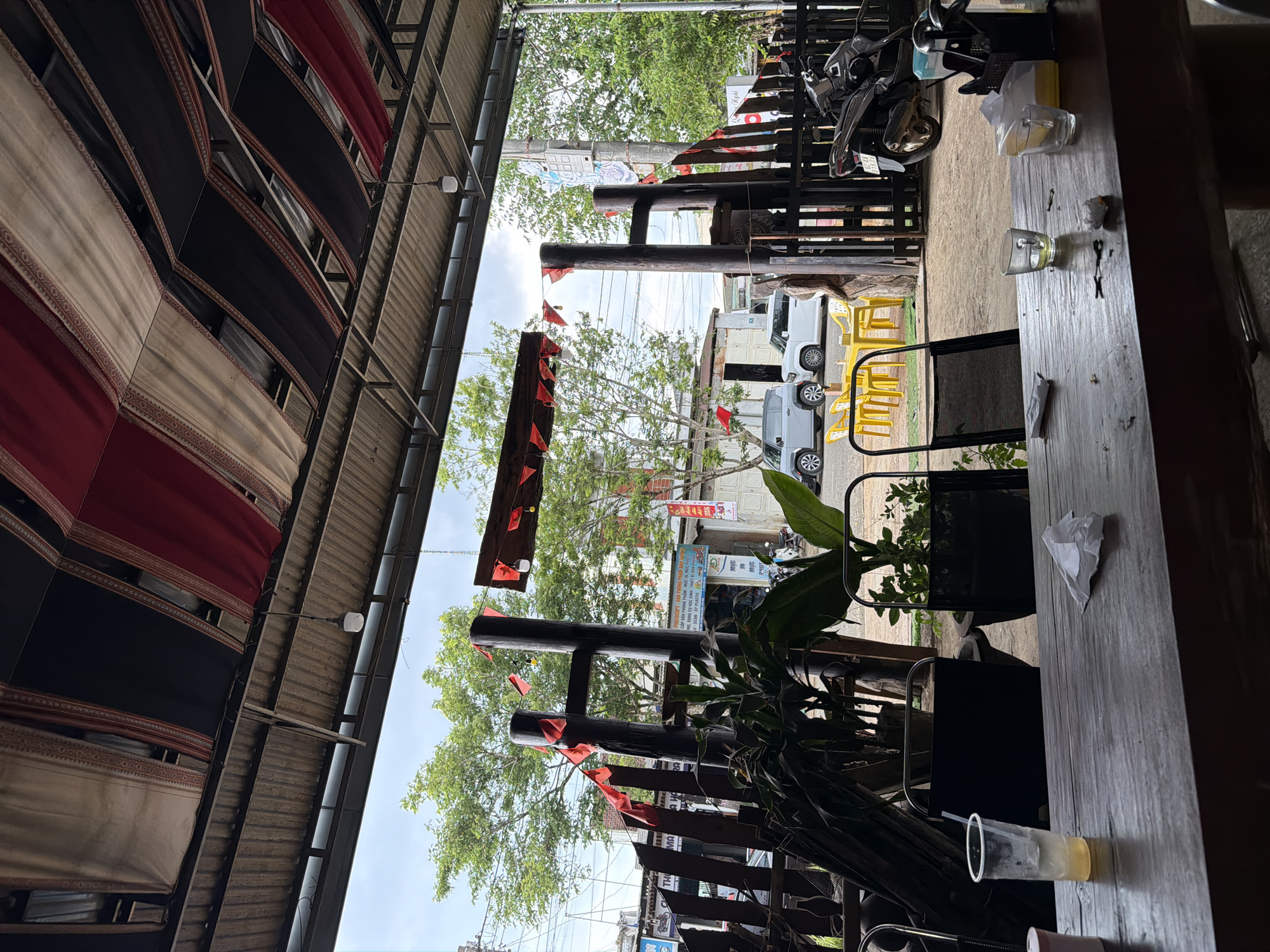 The image depicts an outdoor or semi-outdoor dining area, likely a cafe or eatery, situated alongside a street in Măng Đen, Vietnam. The scene is captured in bright daylight, suggesting midday or early afternoon, with a partly cloudy sky visible.
In the foreground, a sturdy wooden table runs horizontally across the frame. On it are several clear glasses, some containing a light yellow liquid (possibly a drink like iced tea or beer), and one with a straw. Crumpled white tissues are also scattered on the table. To the left of the table, a black metal chair with a dark fabric seat is visible, and a leafy green plant in a pot sits nearby.
The midground features dark wooden posts forming part of the establishment's structure, possibly supporting an awning or roof. A string of small, triangular red flags is draped between these posts, adding a festive or decorative touch. Beyond this, the view opens to a bustling street scene.
The background reveals a street with several white vehicles, including what appear to be vans, and a motorcycle parked on the pavement. Yellow plastic chairs are stacked or arranged near the vehicles. Green trees with lush foliage line the street, partially obscuring buildings in the distance. A power pole with some wiring is also visible.
Overhead, a corrugated metal roof provides shelter for the dining area. Beneath this roof, various lengths of fabric in red, black, and cream, adorned with patterned borders, are draped and suspended, likely serving as decorative elements or to provide additional shade.
Visible text on signs in the background includes Vietnamese phrases such as "TOCOPY VAN", "CẤP VĂN PH", "SCAN ÉP PLASTIC", "AD", "MỰC", "Ngãi", and "THU", suggesting a nearby business like a copy center or stationery shop. No people are visible in the frame, giving the impression of a quiet moment in an otherwise active setting.