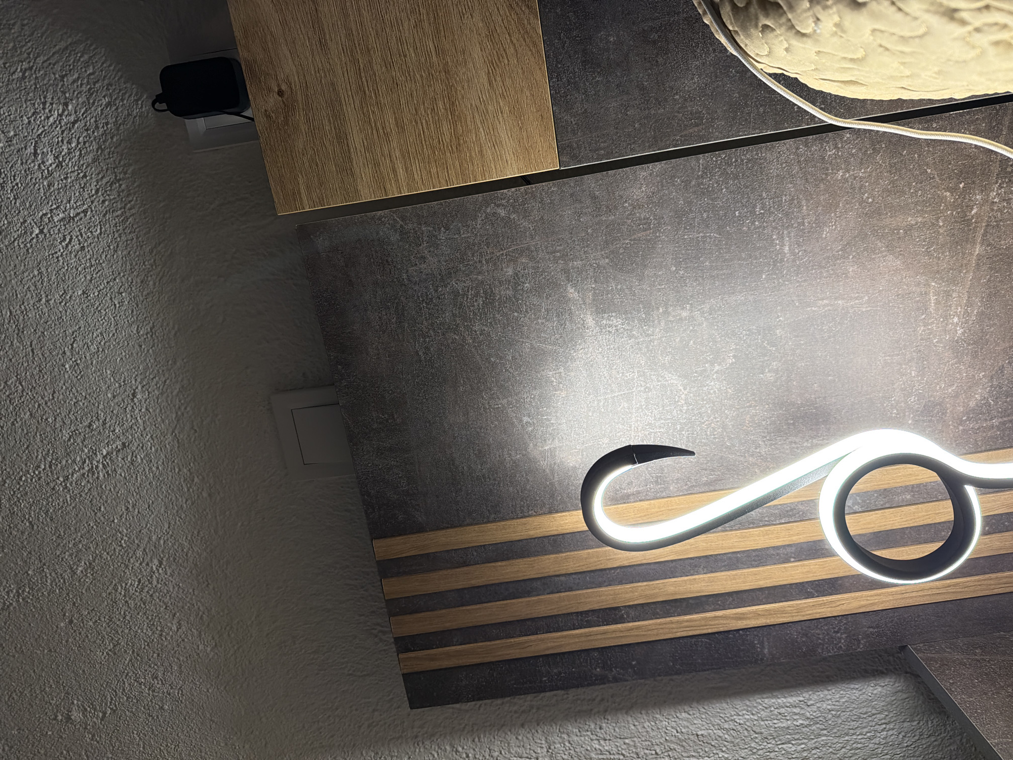 The image shows a close-up interior shot of a room, likely a modern living space or bedroom. The foreground features a neon light fixture, bent into a stylized, abstract shape, possibly resembling a swan or a cursive letter. The light is bright white and casts a glow on the surrounding surfaces. Behind the light, a dark, textured wall panel is visible, with vertical wooden slats interspersed. To the right, a section of a wooden surface with a prominent grain pattern is present, and further to the right, a portion of a textured object, possibly a sculpture or a decorative item, is partially visible. In the upper right corner, a black power adapter is plugged into a wall outlet. The ceiling is textured and white, with a visible light fixture or vent. The overall impression is one of contemporary interior design. There is no indication of the city of Zmeitsa, Bulgaria, in the image.