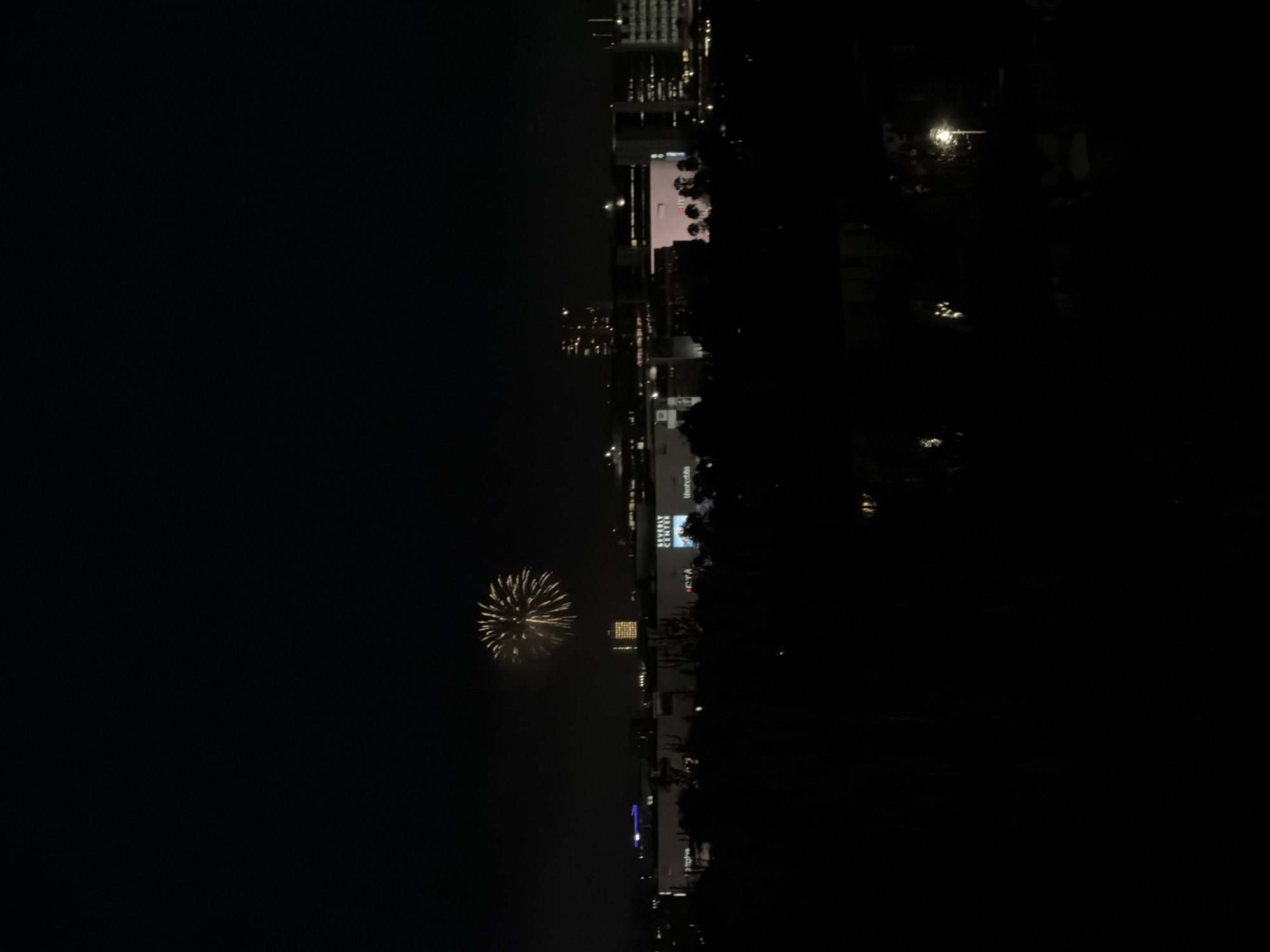 A single, yellow firework bursts in the night sky over the Beverly Center in West Hollywood, California. The cityscape is visible in the background, with the illuminated mall complex taking center stage. Trees in the foreground are silhouetted against the dark night, and a streetlamp glows in the lower right corner of the frame.