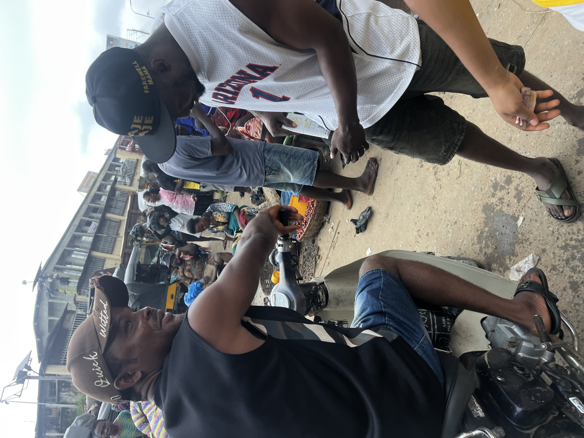 In Nkpor, Nigeria, two men are positioned near a marketplace, one seated on a motorcycle and the other standing. The man on the motorcycle wears a black sleeveless shirt and denim shorts, with a brown baseball cap that reads "Quick witted." The standing man is dressed in a white sleeveless basketball jersey with "ARIZONA" and the number "1" visible, and dark denim shorts. He holds a white packet in his hands. Various individuals are scattered throughout the background, some carrying goods. Structures suggesting market stalls and buildings are visible in the distance, and the ground appears to be a dirt or asphalt surface.