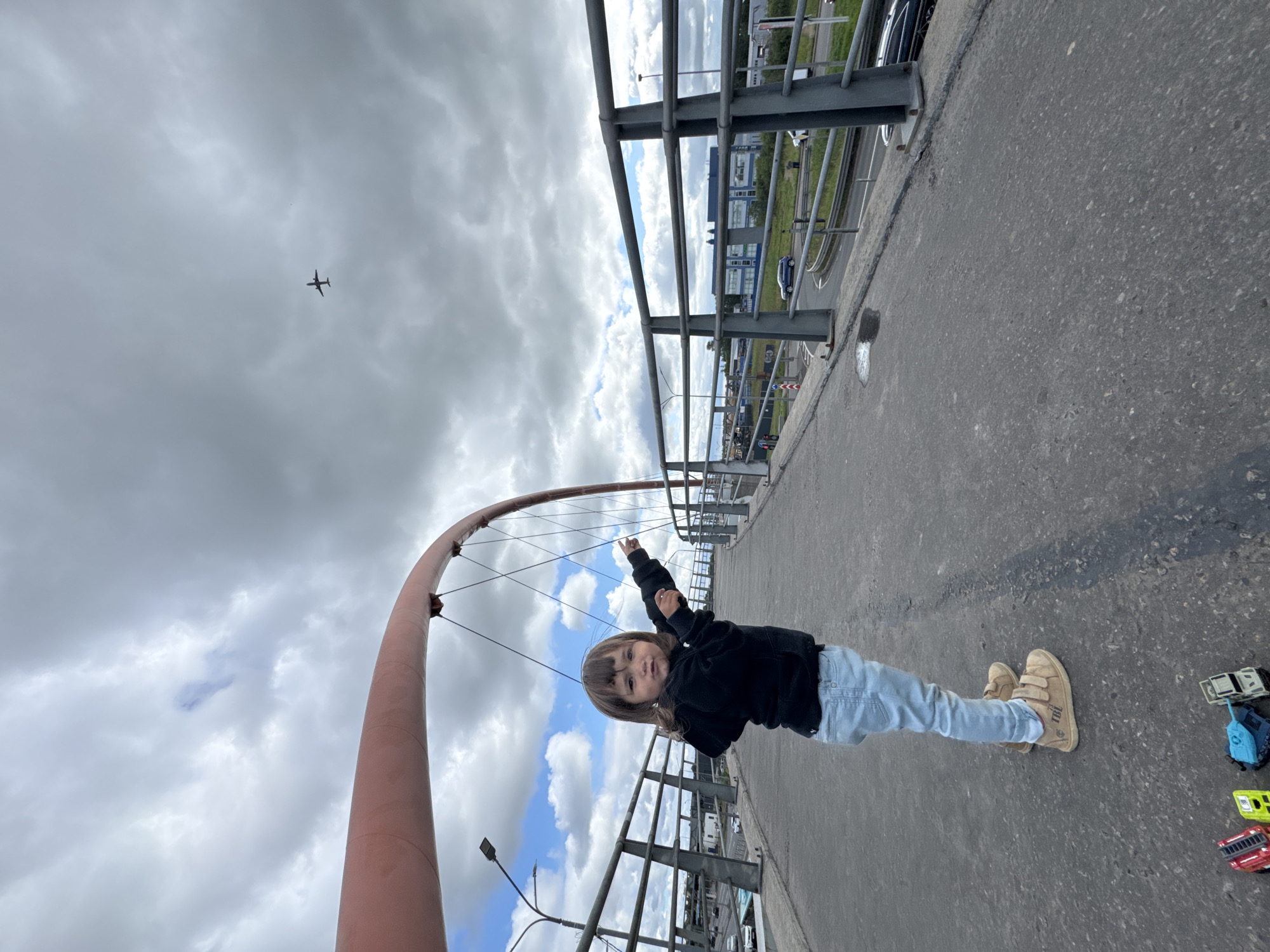 A young child stands on a bridge, pointing at an airplane flying in the cloudy sky. The child is wearing a black hoodie, light blue jeans, and beige sneakers. In the foreground, several toy vehicles, including a red and white truck, a green vehicle, a blue tank, and a gray vehicle, are lined up on the pavement. The bridge has a red arched structure overhead and metal railings along the sides. Below the bridge, a multi-lane road with cars can be seen. The overall environment suggests an urban setting with a focus on the child's observation of the aircraft.