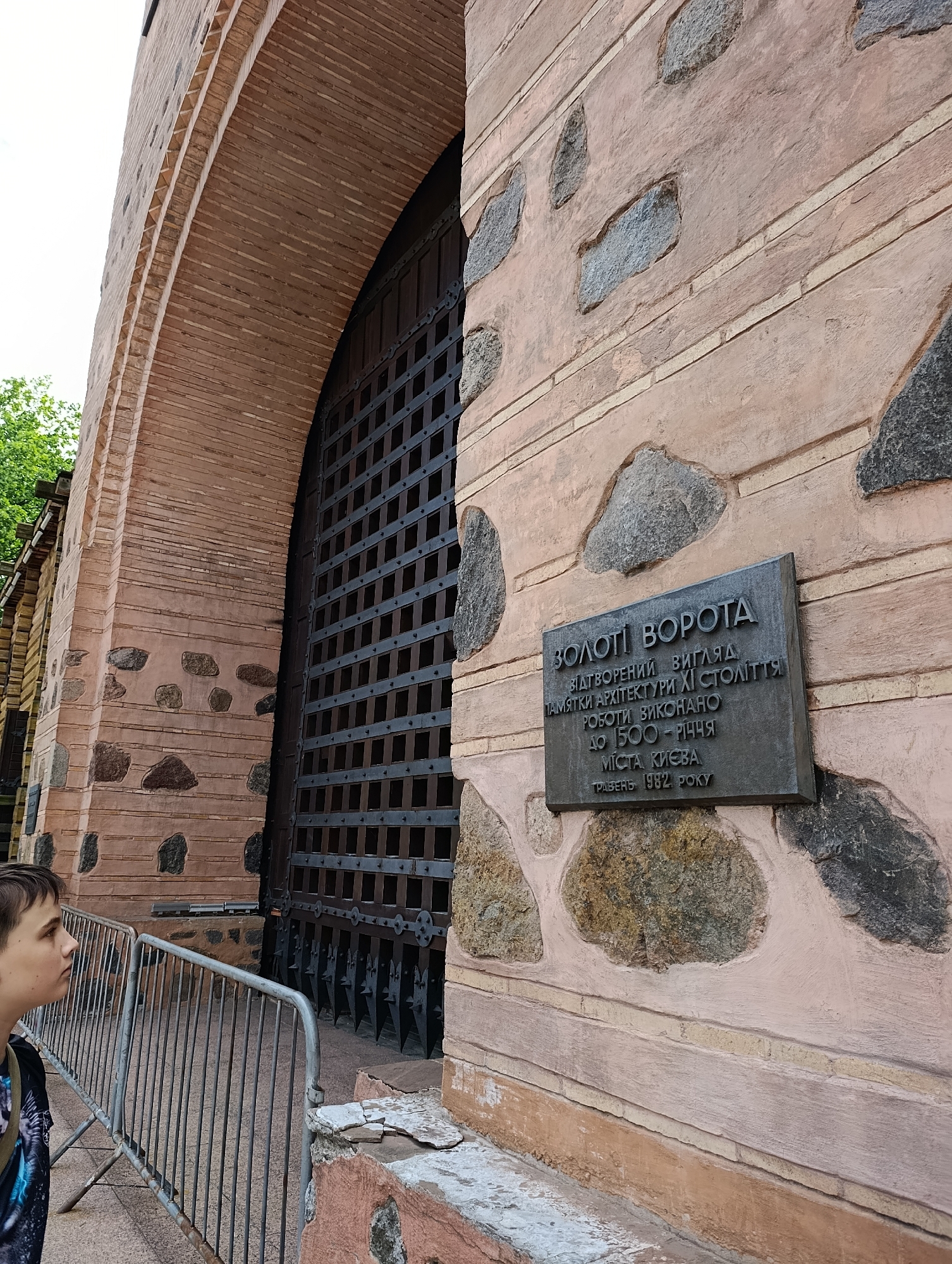 An outdoor, eye-level, medium shot depicts the Golden Gate in Kyiv, Ukraine. The gate is a large, arched structure made of reddish-brown bricks and large, rough-hewn stones. A tall, dark metal gate with a grid pattern is visible within the archway. A plaque is mounted on the wall to the right of the gate, bearing text in Ukrainian. A young boy is partially visible on the left side of the frame, looking towards the gate. A metal barrier is in the foreground, separating the viewer from the gate. The sky is overcast.