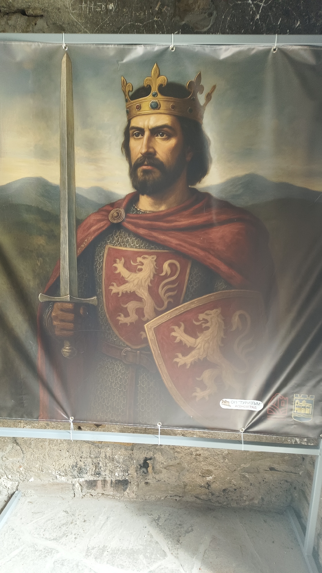 The image depicts a banner or large print of a painting, likely displayed within a historical or touristic site. The painting portrays a man with a beard and dark hair, wearing a golden crown, a red cloak, and chainmail. He holds a sword in his right hand. Two shields are visible on his chest, both bearing the image of a lion. The backdrop of the painting shows a mountainous landscape. The banner itself is suspended by hooks from a metal bar. The background includes a stone wall and what appears to be the inside of a stone structure or room. A logo is visible on the lower right corner of the banner, that says "ОП "ТУРИЗЪМ" АСЕНОВГРАД". This logo suggests that the location is in or associated with Asenovgrad, Bulgaria.