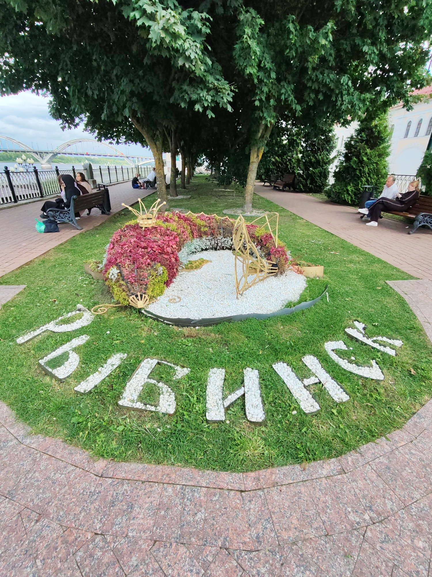 The image depicts a park-like setting with a prominent floral arrangement in the shape of a mythical creature, possibly a dragon or a fish, adorned with a golden crown. The creature is positioned on a bed of white pebbles, surrounded by lush green grass. In front of the floral arrangement, large, white stone letters spell out "РЫБИНСК" (Rybinsk) on the grass. Several people are visible in the background, some sitting on park benches under the shade of trees, while others are walking along a paved pathway. A bridge with multiple arches spans a body of water in the distance. The overall environment suggests a public space, likely a park or promenade in the city of Rybinsk, Russia.