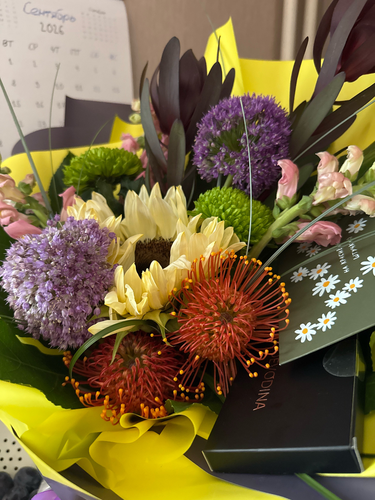 A floral arrangement featuring sunflowers, purple alliums, pincushion proteas, green chrysanthemums, and pink snapdragons is displayed prominently. The bouquet is wrapped in bright yellow paper and rests on a dark surface, possibly a box. A calendar with "Сентябрь 2026" (September 2026) written in Cyrillic is visible in the background. A dark green card adorned with small white daisy illustrations and Cyrillic text is placed within the bouquet. The scene appears to be indoors, with a suggestion of an office or domestic setting based on the calendar and general lighting. The location in the city of Захарьино, Russia, is noted, though no specific landmarks or contextual clues within the image directly confirm this.