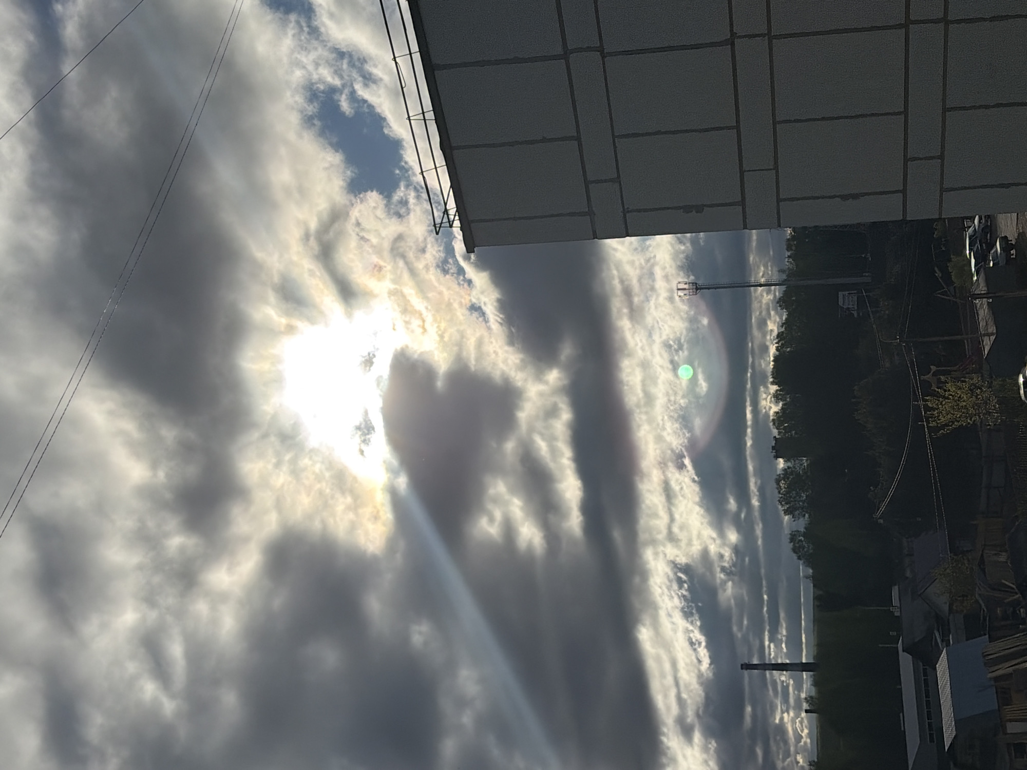 The image shows a view from a high vantage point, possibly a building in the city of Chaikovsky, Russia. The sky is filled with a mix of dark and light clouds, with the sun partially obscured. Sun rays are visible, emanating from behind the clouds. A building is partially visible at the top of the image. In the distance, a line of trees can be seen, along with parts of the urban environment, including buildings and a tall structure with a slender cylindrical shape. There are signs of construction or industrial activity. Power lines are visible.