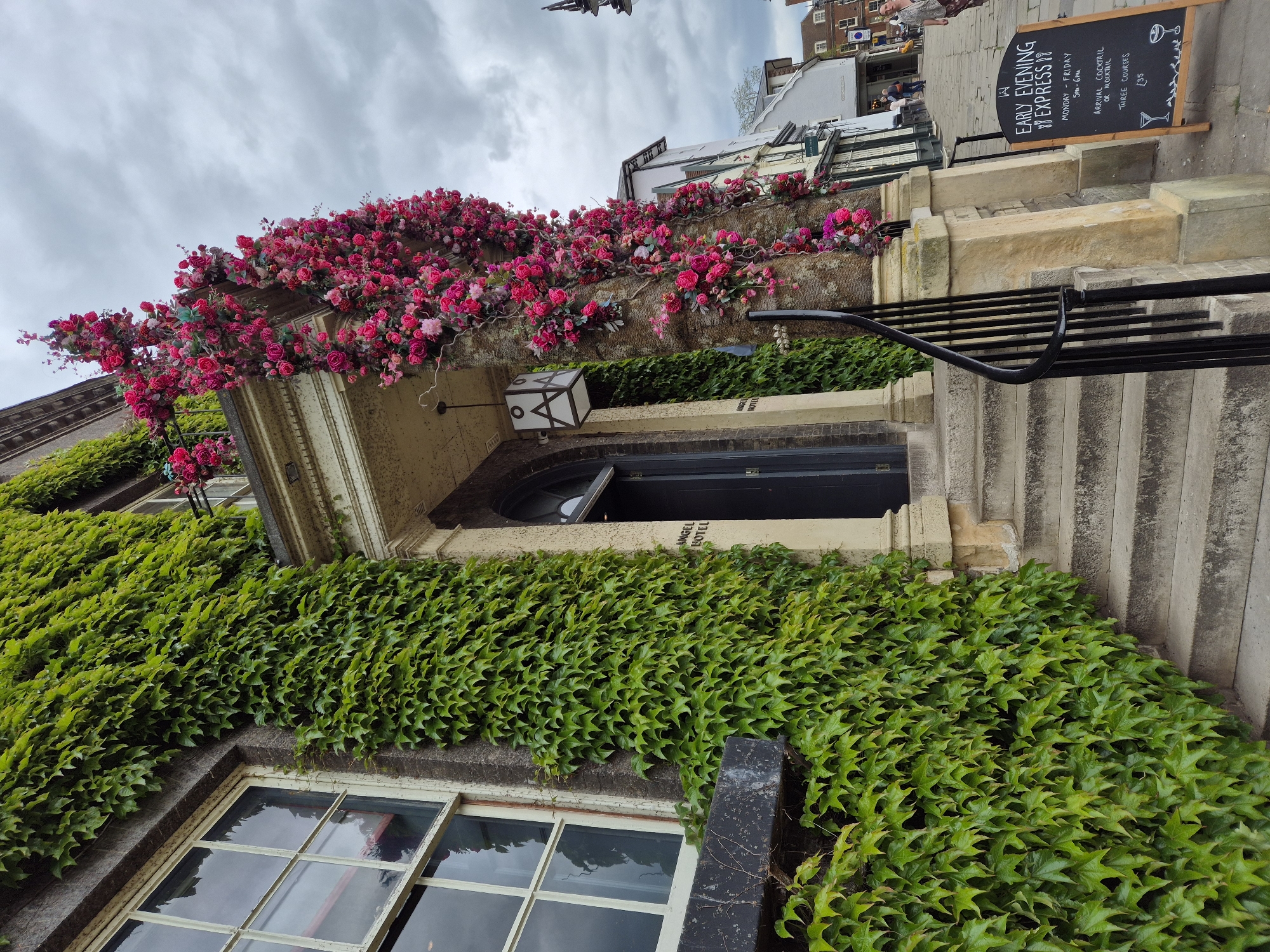 The image captures the exterior of a building, likely a hotel, adorned with lush greenery. The bottom portion of the facade is covered in ivy, partially obscuring a window. Above, a doorway is also framed by ivy, with pink floral arrangements decorating the ledge above it. The entrance steps are visible on the right-hand side, with a metal railing. A black chalkboard sign stands beside the steps. The chalkboard reads "Early Evening Express!" and details a Monday to Friday special for £35. It offers an arrival cocktail or mocktail. The location is West Suffolk, United Kingdom, as indicated by the text "Angel Hotel" above the entrance. The sky is overcast and cloudy.