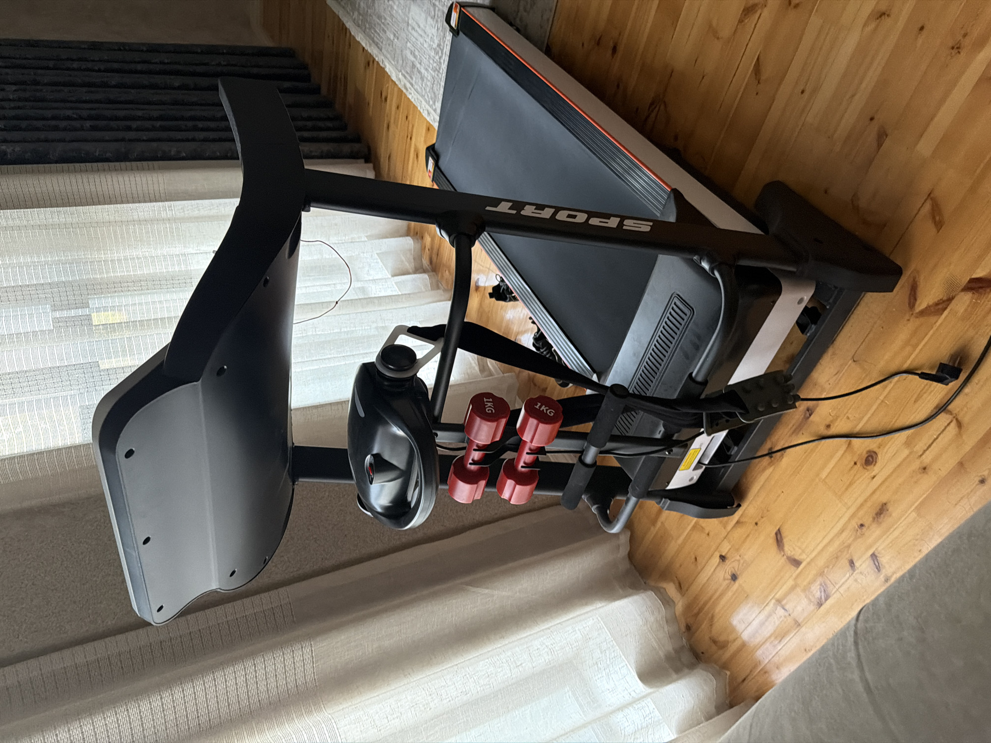 A black and white treadmill is positioned on a wooden floor in what appears to be a home or indoor space. The treadmill has a black running belt with orange trim, and a console at the front. Attached to the treadmill's frame are two red 1kg dumbbells. Sheer white curtains are visible in the background. The image is taken from a slightly elevated angle, looking down at the treadmill. The lighting suggests it is daytime, with natural light filtering through the curtains. There are no people or animals present in the image. The environment appears clean and organized. The provided location "Sabirabad, Azerbaijan" is not visually supported by the image content.