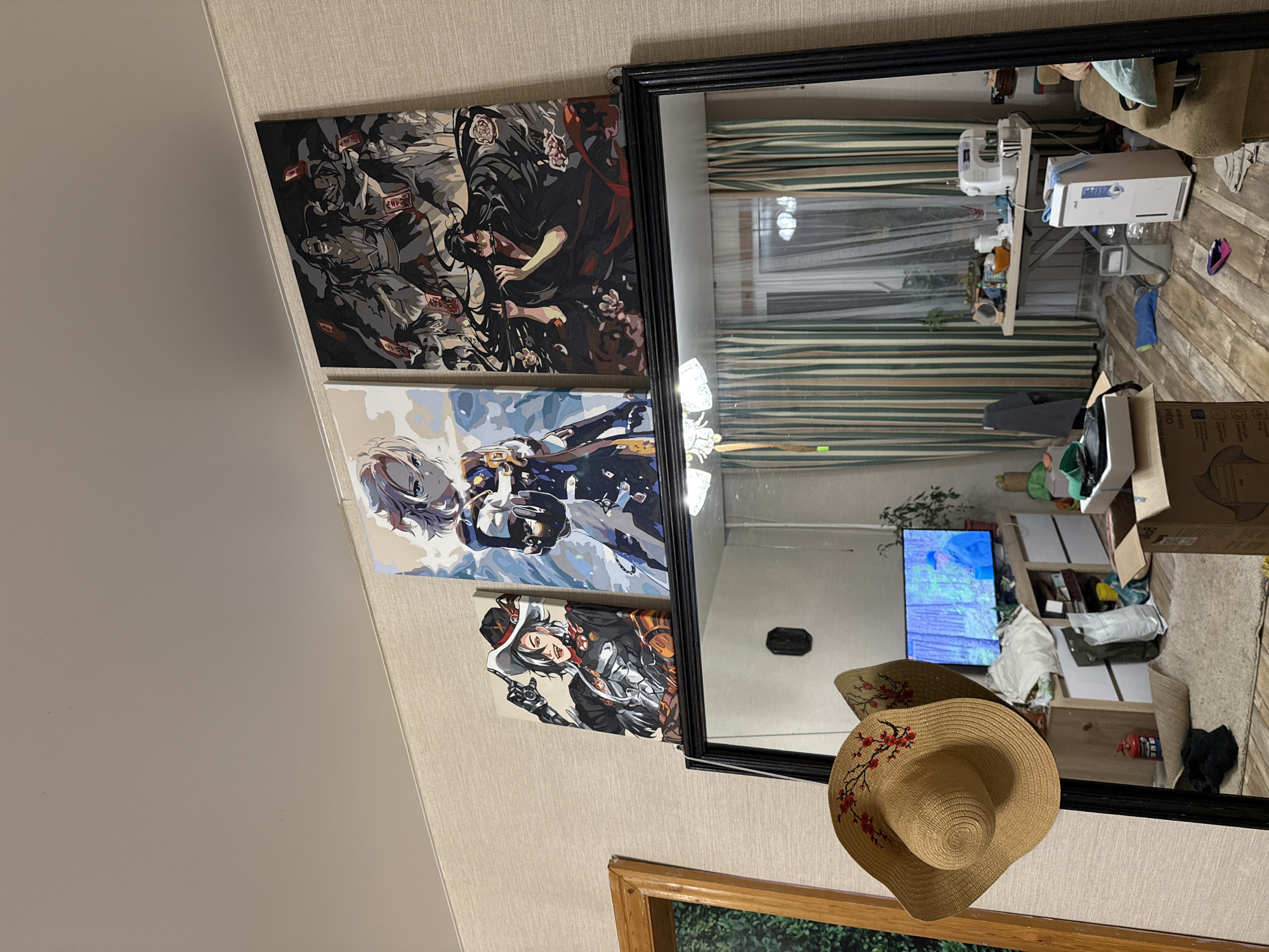 The image depicts an indoor scene, likely a living room, with a large mirror dominating the foreground and reflecting the opposite side of the room. Above the mirror, three framed anime-style artworks are displayed on a textured wallpapered wall. The artwork on the left shows a male character in a cowboy hat and attire, the center artwork features a female character in a blue and white outfit, and the right artwork displays multiple figures in dark clothing.
The mirror's reflection reveals a cluttered living space. A television screen is visible, showing an image of a forest with a person. In the reflection, a sewing machine sits on a table against a wall with striped curtains. Various household items, including boxes, bags, and a dehumidifier, are scattered on the wooden floor and on shelves. A straw hat with red floral decorations is hanging on the left side of the mirror. The overall impression is one of a lived-in, somewhat disorganized domestic environment. The location is identified as Sovetskiy, Russia, suggesting a specific geographical context.