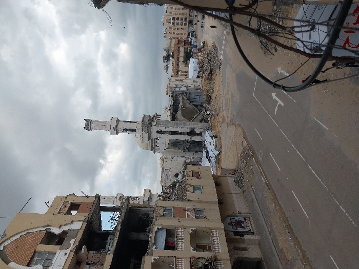 This image depicts a heavily damaged urban landscape in Khan Yunis, Palestinian Territory, under an overcast sky. In the mid-ground, a severely damaged mosque with a partially collapsed minaret and damaged dome stands amidst piles of rubble. To the left, a multi-story building shows extensive structural damage, with entire sections of walls missing, exposing interior rooms, and debris scattered below. Balconies on this building show some items, including what appears to be laundry. A large mural or poster is visible on its lower exterior wall.
An asphalt road with white lane markings, including right-turn and straight arrows, traverses the foreground, separating the mosque and the damaged building on the left. Extensive debris fields, consisting of concrete, metal, and other building materials, are spread across the ground, particularly around the mosque and the damaged buildings. In the far background, further damaged structures are visible. Several indistinct figures are seen walking on a street or path in the far-right background. Prominent wires and cables, some appearing detached, are visible in the immediate foreground on the right, partially obscuring the view.