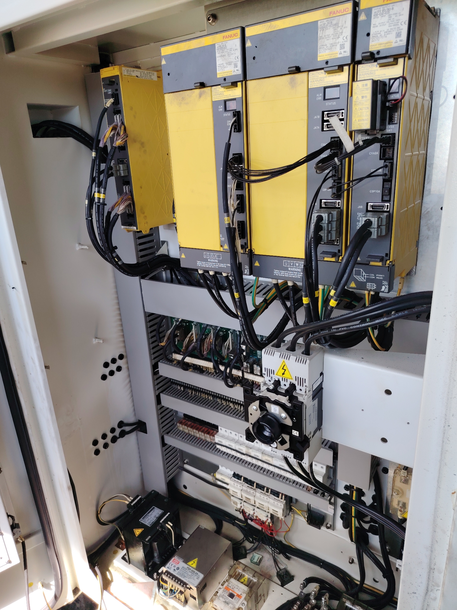 This media file shows the interior of an industrial electrical control cabinet, likely for automation machinery. The cabinet is filled with electronic components, including several yellow and gray modules labeled "FANUC," which are likely servo drives or controllers. Numerous cables, both black and multicolored, are neatly routed and connected to these modules and other components within the cabinet. Some of the modules have warning labels and status indicators.
Below the FANUC modules, there is a rack of smaller components, including what appear to be terminal blocks, circuit breakers, and possibly relays. A prominent component with a yellow triangle warning symbol and a silver dial is mounted on the upper part of this rack. Towards the bottom of the cabinet, there are additional power supply units and transformers, also bearing warning labels. The overall impression is one of complex, organized electrical infrastructure.
There are no people visible in the image. The lighting suggests an indoor setting, possibly a factory floor or a maintenance area. The specific location within Noviy, Russia, is not visually indicated beyond the general context of industrial equipment. The time of day and weather are not discernible from the image. Visible text includes brand names like "FANUC" and "CE," as well as warning messages and technical labels on the components. There is also text on cables, such as "FIOI ZHAO TAI PVC CONTROL WIRE CABLE 14mm".