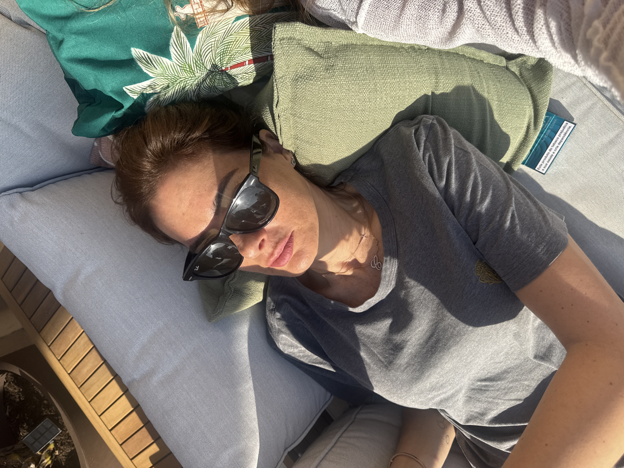 A woman with brown hair and sunglasses lies on a gray cushioned lounge chair with green pillows in an outdoor setting. She wears a gray t-shirt and a delicate necklace. A turquoise IQOS ILUMA package, marked with a health warning in Portuguese, is visible near the pillows. The location, though not explicitly stated, is implied by the Portuguese text to be in Portugal. The precise city, Vila Nova de Gaia, cannot be confirmed visually.
