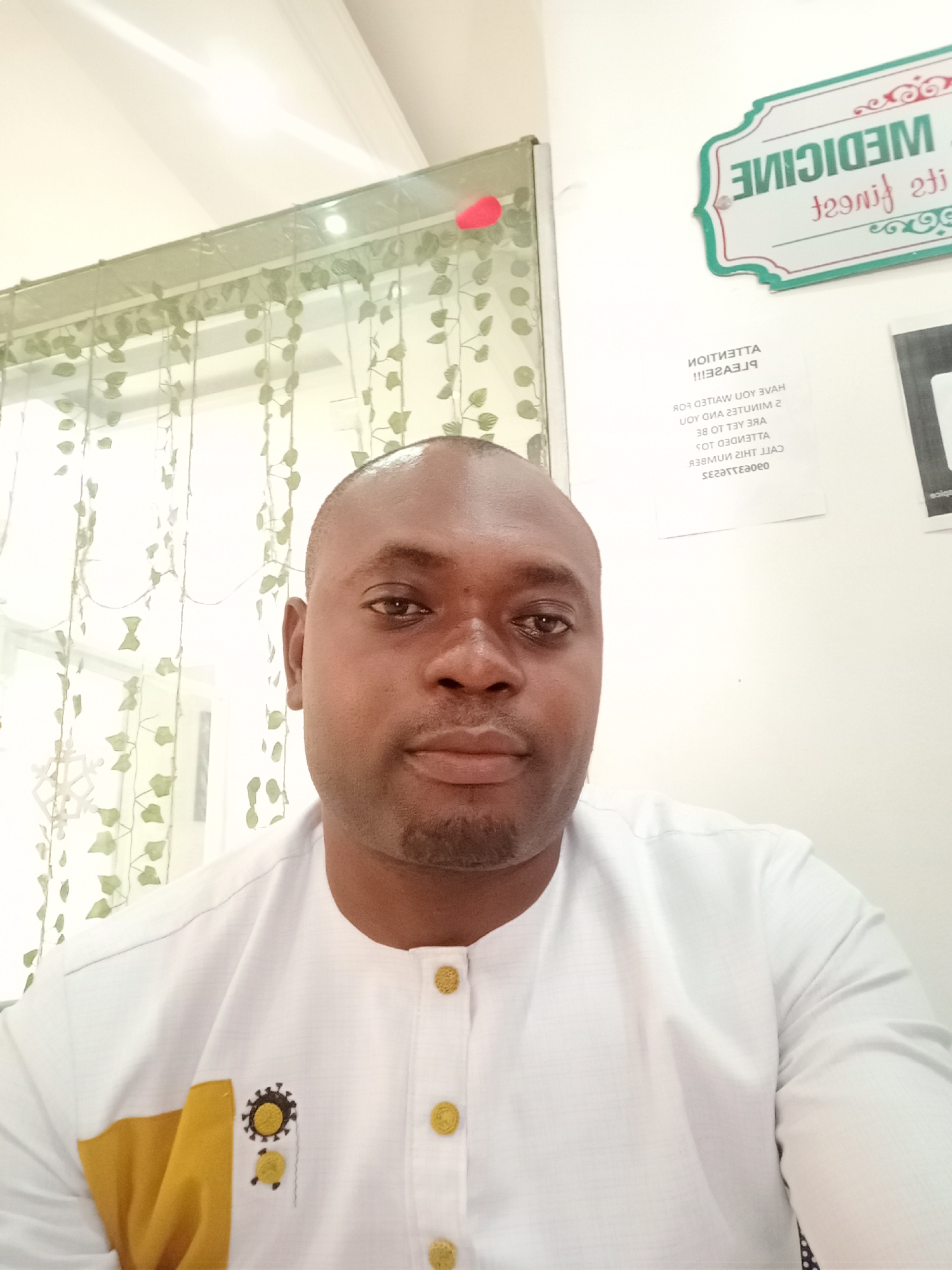 A medium close-up selfie shows a bald man, likely in Gwarinpa, Nigeria, looking directly at the camera with a neutral expression. He has a light beard/stubble. He is wearing a white collared shirt with gold-colored buttons running down the front placket. The left side of his chest features a design with a yellow block and black/gold embroidery that resembles a gear or virus icon.
The background is indoors, possibly an office or waiting area. On the left, light green sheer curtains with leaf patterns are visible. To the right of the man, a white wall displays several signs. The most prominent sign, at the top right, reads "MEDICINE its finest" in reverse, framed with a green border and red outline. Below this, another sign with reversed text appears to be a notice. Partially legible sections, reading backward, include "ATTENTION PLEASE!!" and phrases like "HAVE YOU WAITED FOR," "23 MINUTES AND YOU ARE TO BE ATTENDED," "CALL THIS NUMBER," followed by a sequence of characters that might be part of a phone number or code. Overhead, bright indoor lighting is visible, reflected in the ceiling. The overall lighting suggests it is daytime.