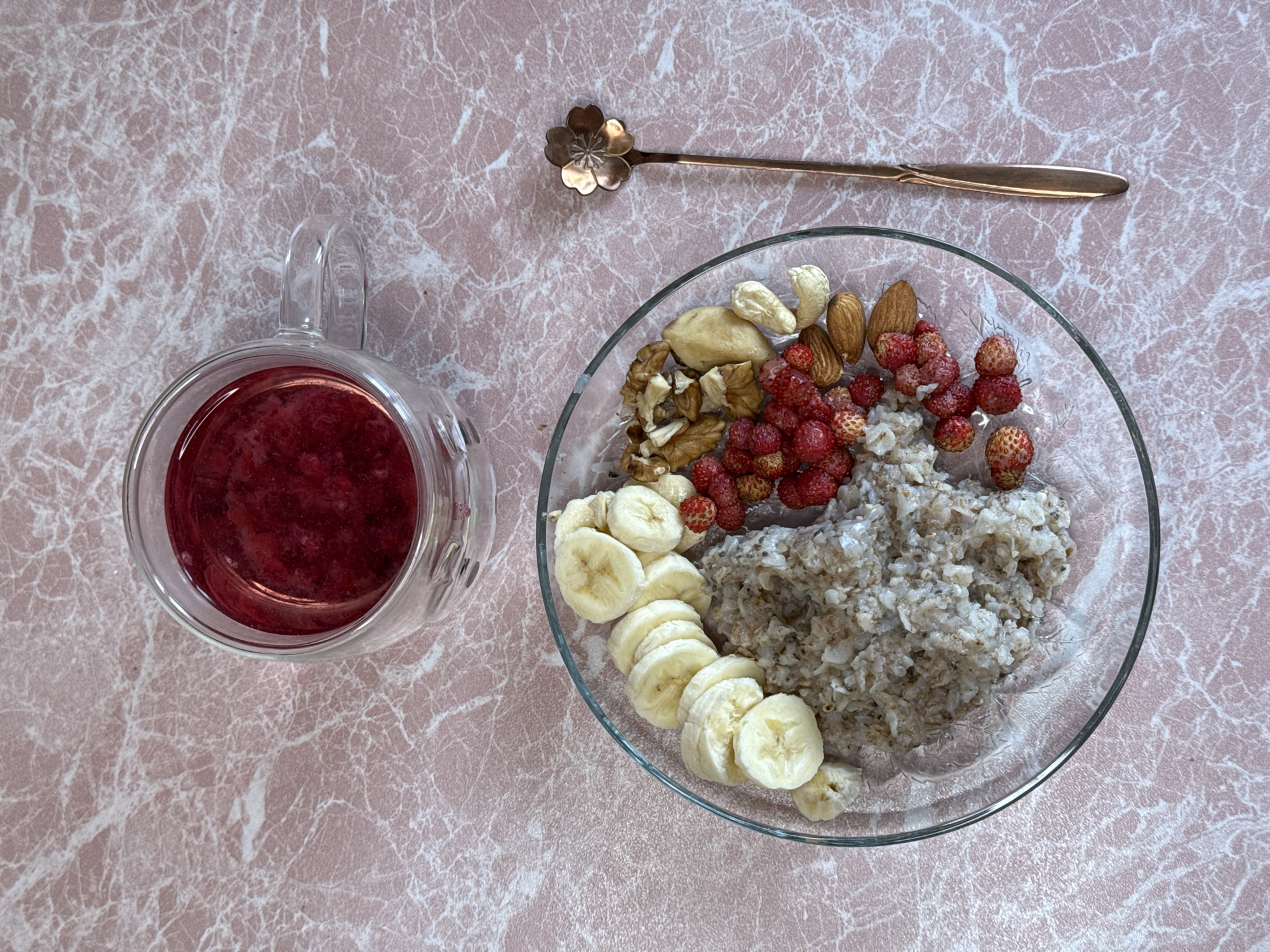 An overhead, eye-level shot shows a clear glass mug filled with a dark red beverage on a pink marble-patterned surface. To the right of the mug is a clear glass plate containing a portion of oatmeal, sliced bananas, small wild strawberries, almonds, cashews, and walnuts. A rose gold spoon with a flower-shaped head lies to the right of the plate. The scene appears to be indoors, likely on a tabletop.