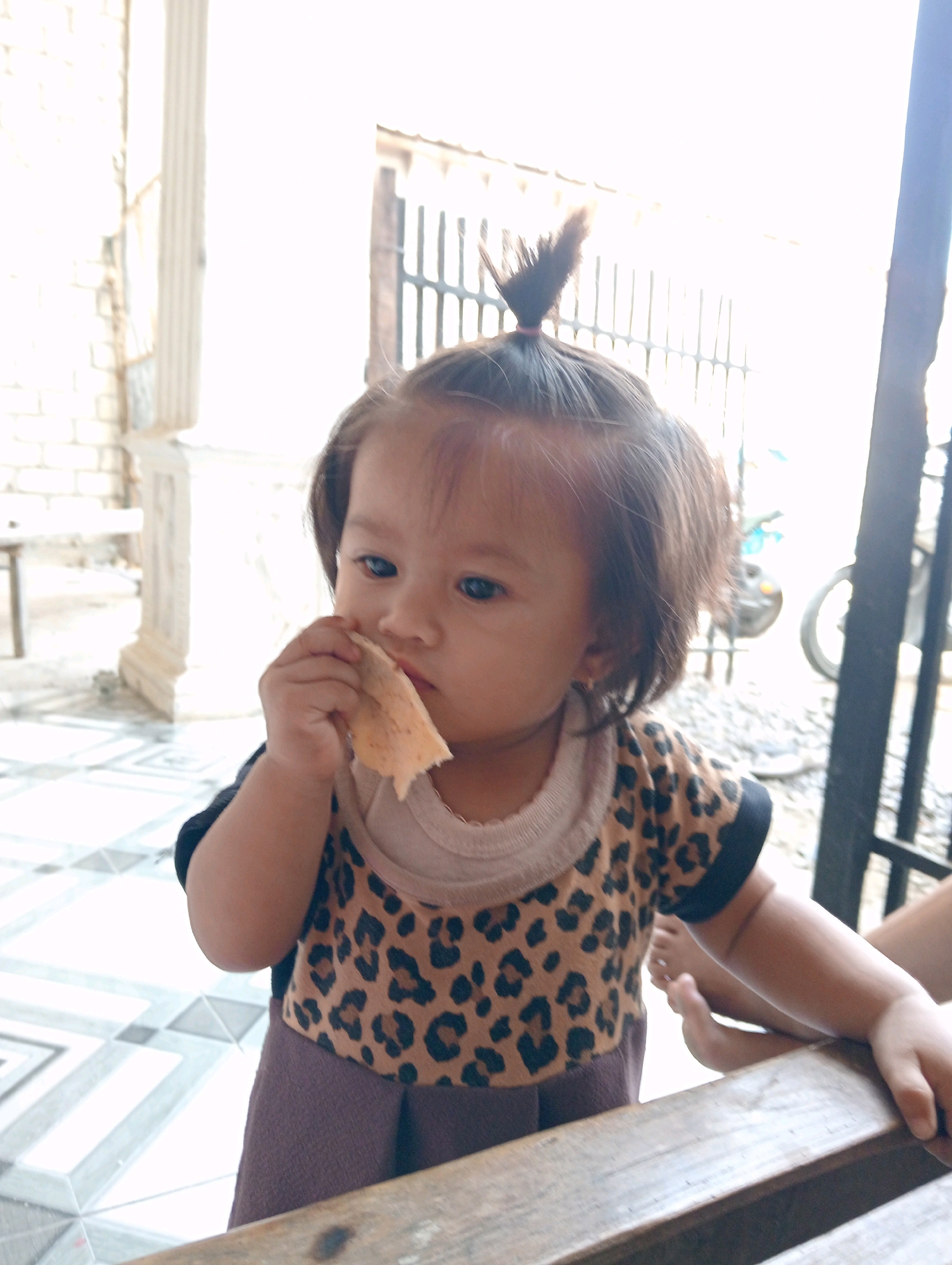 A young child, appearing to be of Asian descent, is captured in a close-up portrait, holding a piece of food to their mouth and chewing. The child has dark hair styled in a small bun on top of their head, and their eyes are focused on the food. They are wearing a leopard-print shirt with a light-colored neckline and a dark-colored trim on the sleeves. Their lower body is clad in a purple skirt. The child is positioned at a wooden table, with their left arm resting on it. In the background, the scene suggests an outdoor or semi-outdoor setting. A brick wall and a white pillar are visible on the left, with a metal gate and a motorcycle in the distance on the right. The ground is tiled with a geometric pattern of white and gray squares. The lighting is bright, indicating daylight. Based on the context provided, this scene is set in the city of Soko, Indonesia.
