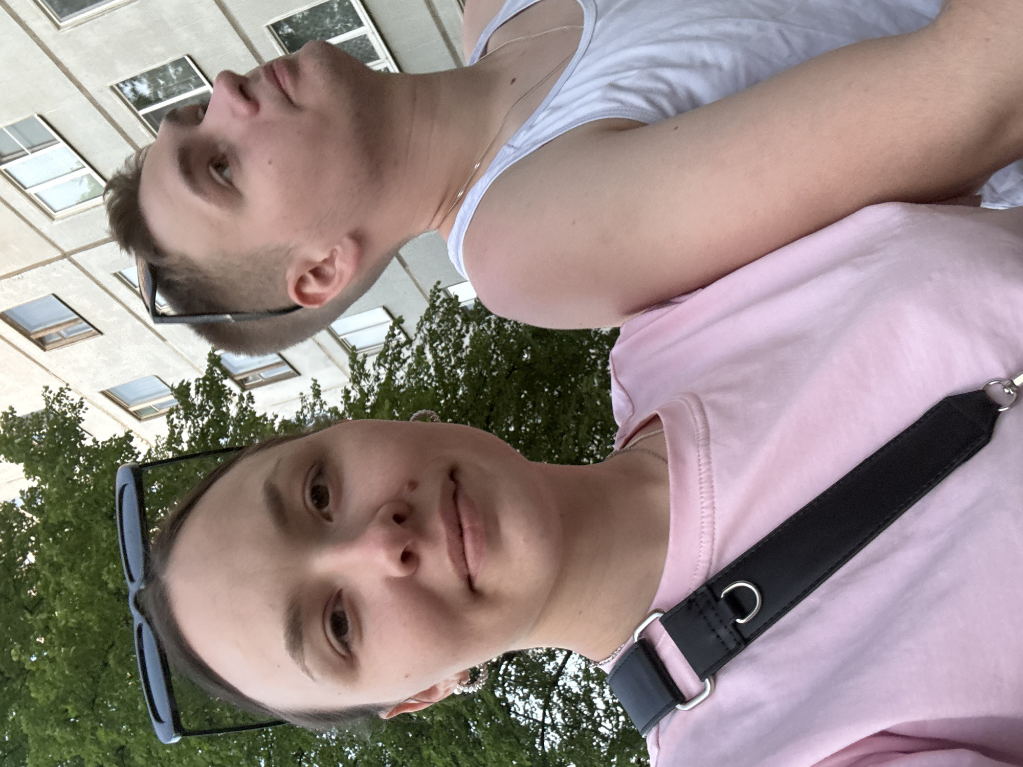 Two individuals are captured in a selfie-style image. The person in the foreground is wearing a pink t-shirt and black-rimmed sunglasses. A black strap is visible across their torso. The person in the background is wearing a white tank top and glasses. Both individuals are fair-skinned. Behind them is a building with multiple windows and trees, suggesting an urban environment. The image was taken in the city of Chaikovsky, Russia.