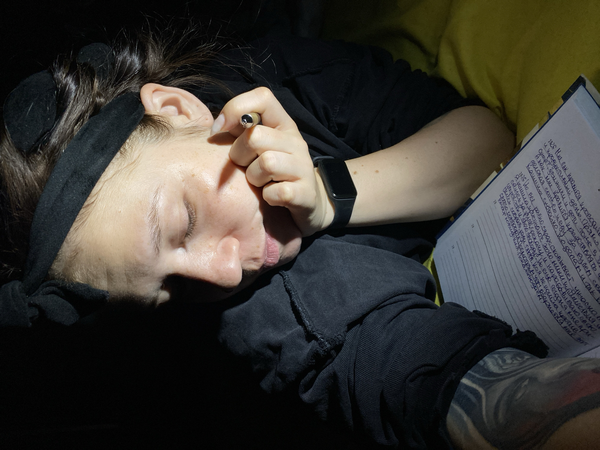 A young woman with closed eyes rests her head on her hand, propped up by her arm. She is wearing a black headband and a black hoodie. A smartwatch is visible on her left wrist, and she is wearing a ring on her right index finger. In front of her is an open notebook with handwritten text in Russian. Her left arm has tattoos. The lighting suggests she is indoors, possibly at night, with a light source illuminating her face and upper body from the side, casting strong shadows. There is no discernible evidence to determine the specific location within the city of Chaikovsky, Russia.