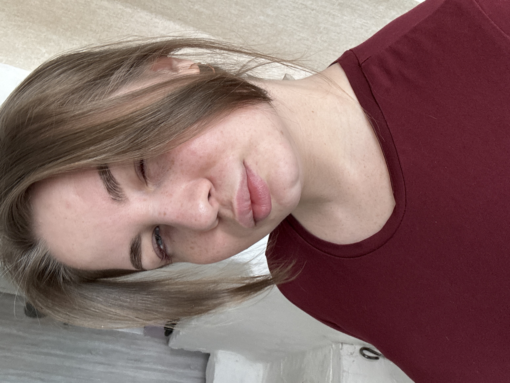 A close-up portrait of a young, fair-skinned woman with freckles, wearing a maroon-colored shirt. She has light brown hair with some strands falling across her face. Her eyes are partially closed, and her lips are pursed. The background appears to be a simple wall or surface. Location is unspecified.