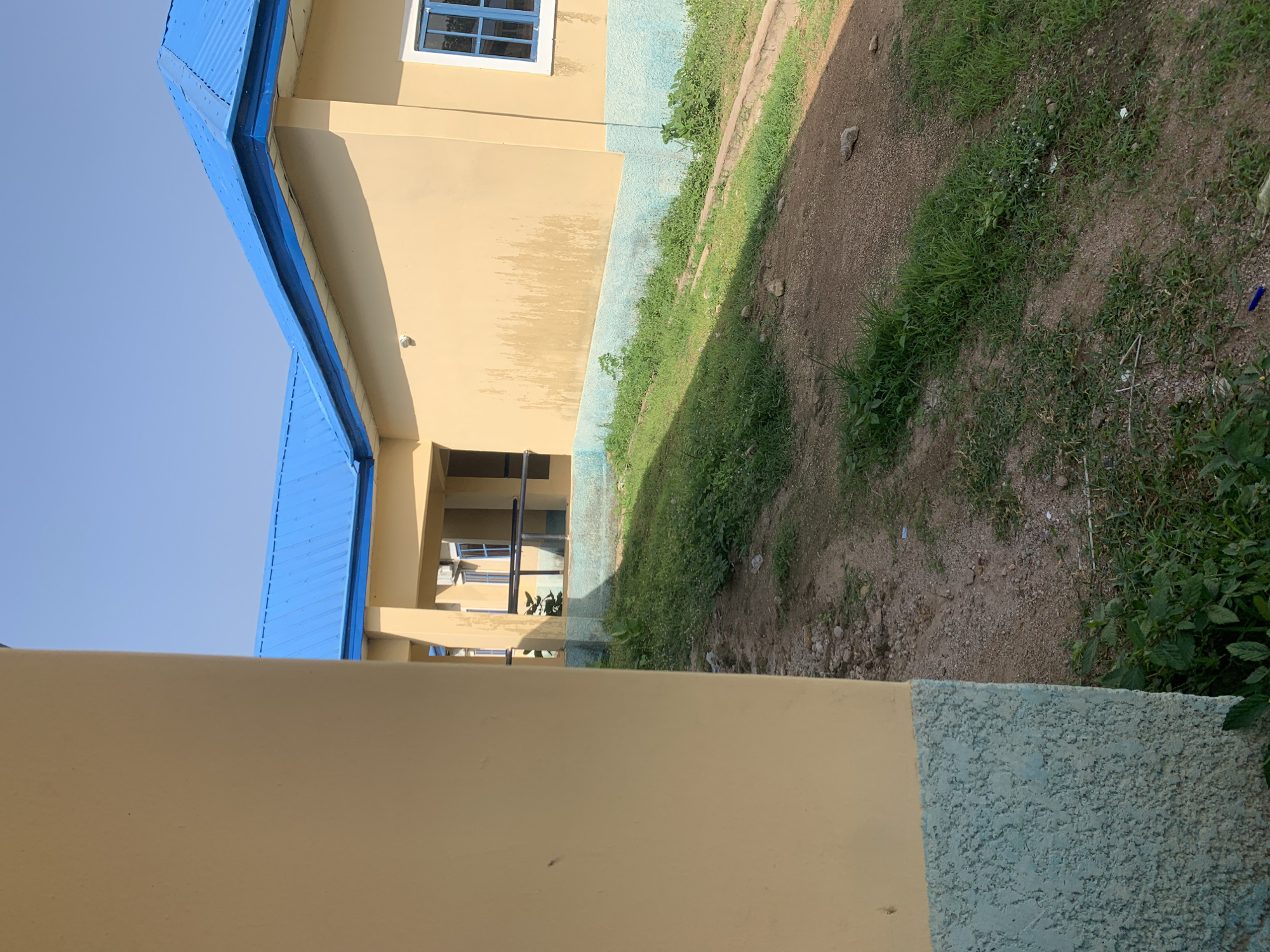 A building with a bright blue, corrugated metal roof stands under a clear blue sky. The walls of the building are painted a pale yellow, with a rough, light blue textured band near the foundation. A window with a blue frame is visible on the right side of the building. The foreground shows a patch of dry, brown earth and sparse green grass, with a few small stones scattered about. The ground slopes downwards from the building. This scene is located in Jalingo, Nigeria.