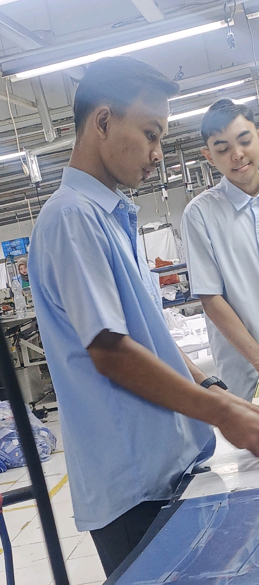 Two young men, likely factory workers, are shown in a brightly lit industrial setting, possibly a textile or garment factory in Salatiga, Indonesia. The man in the foreground, facing right, wears a light blue collared shirt. His skin is darker and he has short black hair. He appears to be engaged in some task, possibly inspecting or handling fabric, as his hands are positioned on a surface. To his right, another young man in a similar light blue shirt is partially visible, facing left and looking downwards, also seemingly occupied with work. The background reveals industrial equipment, shelving with stacked white materials, and overhead lighting fixtures. The overall environment suggests a production or assembly line.