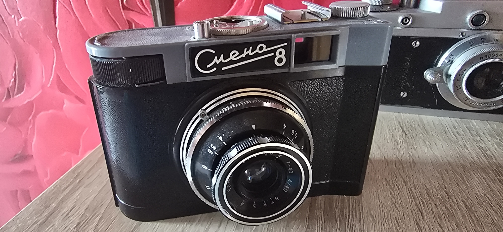 This media file features two vintage film cameras, likely in a domestic setting in Gostagayevskaya, Russia. The primary subject is a black and gray "Smena 8" camera, positioned in the foreground. It has a distinctive silver lens assembly with aperture markings visible, including "5.6" and "8". The camera also has a silver winding knob and a chrome viewfinder. The "Smena 8" text is prominently displayed in white cursive script on the camera's body.
To the right and slightly behind the Smena 8 is another vintage camera, appearing to be silver and black. Some text is visible on its lens assembly, including "N80384" and "F=5см".
The cameras are placed on a wooden surface, possibly a table, which has a light brown, grainy texture. The background features a vibrant, textured pink surface with a floral or petal-like pattern. The lighting suggests an indoor setting, possibly during the daytime, with no specific weather cues. The overall impression is one of a still-life composition focusing on retro photography equipment.