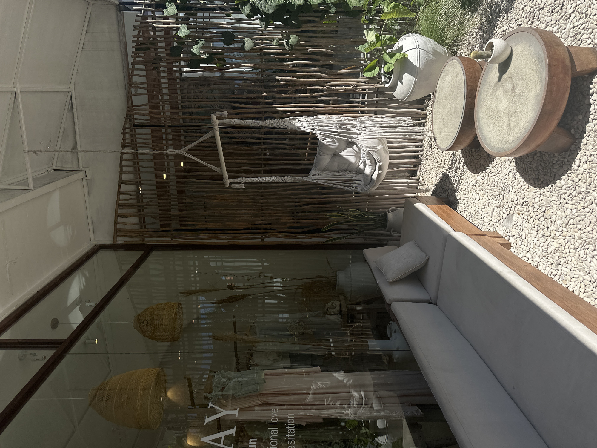 The image depicts a seating area in what appears to be an outdoor or semi-outdoor space in North Kuta, Indonesia. Visible elements include a woven wooden fence, various decorative items, a hammock-style swing, a long white sofa with cushions, and stone landscaping. The scene is viewed through a glass storefront, reflecting light fixtures and potentially merchandise inside the establishment. There are no people visibly present.