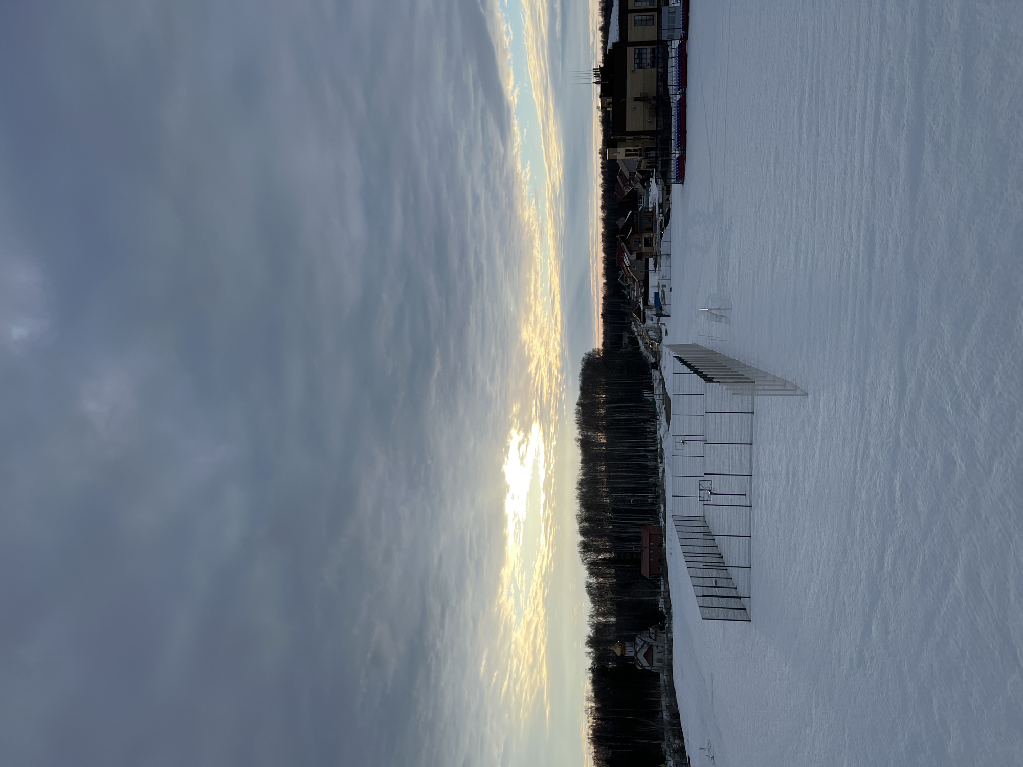 A wide-angle photograph depicts a snow-covered landscape in Noviy, Russia, under an overcast sky at either sunrise or sunset. A bright, golden-yellow sunlight patch breaks through the grey-blue cloud cover on the horizon. The foreground and midground feature a vast expanse of snow, exhibiting minor undulations. Within this area, a fenced sports court, likely for basketball, is visible, containing two basketball hoops with backboards, all covered in snow. A dense line of bare deciduous trees forms a dark horizontal band behind the court. To the left of the tree line, a small Russian Orthodox church with a light facade, red roof details, and a golden onion dome topped with a cross is present. To the right of the church and near the trees stands a small, red, single-story building. The right background reveals a cluster of residential buildings, including several houses and a larger, modern structure with a flat roof and roof-mounted antennae. A roadway and a seating structure with red and blue elements are also visible in the residential zone. No individuals, activities, or interactions are depicted.