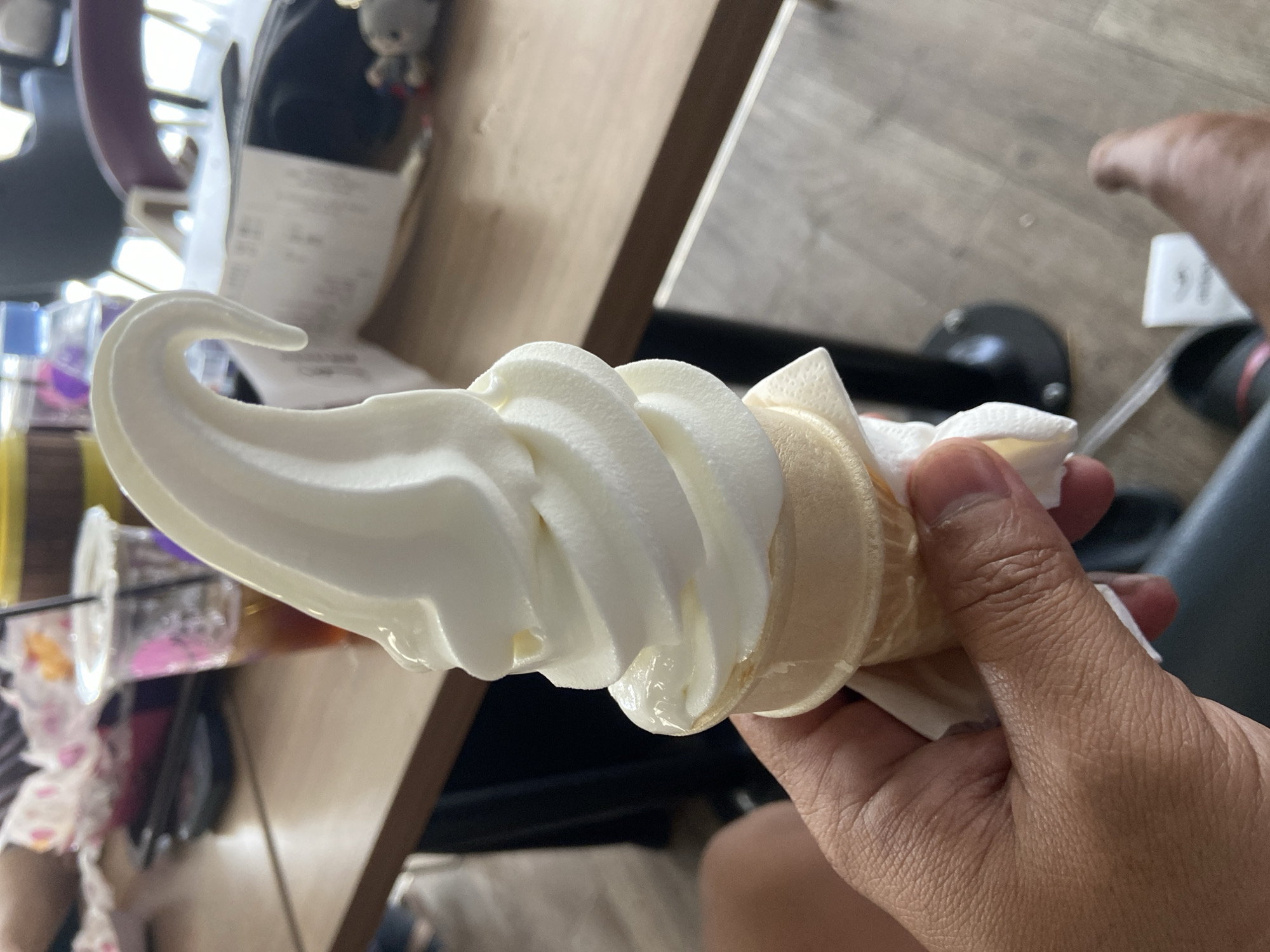 A hand is holding a vanilla soft-serve ice cream cone. The ice cream is a creamy white color and has a swirl pattern. The cone is a light brown color and has a paper wrapper around the bottom. In the background, there is a table with a cup of iced tea, a bottle of water, and a receipt. The scene appears to be in an indoor setting, possibly a restaurant or cafe. The image does not provide any specific information about the city of Bago, Indonesia.