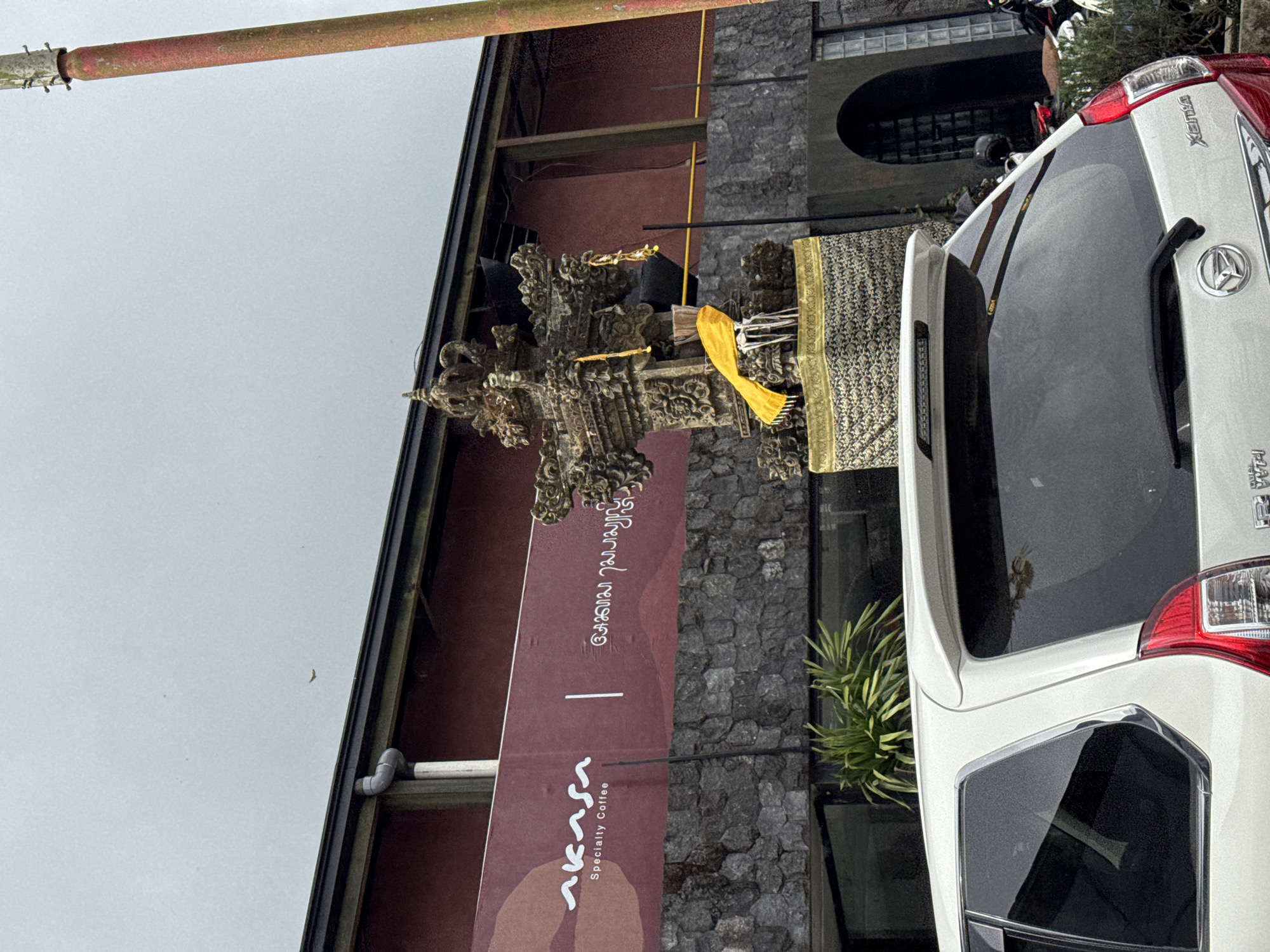 In Kintamani, Indonesia, a white car is parked in front of a building with a dark red facade and a stone base. A decorative stone structure, adorned with yellow fabric, stands near the entrance. The sky is overcast. The building has a sign that reads "Akasa Specialty Coffee."