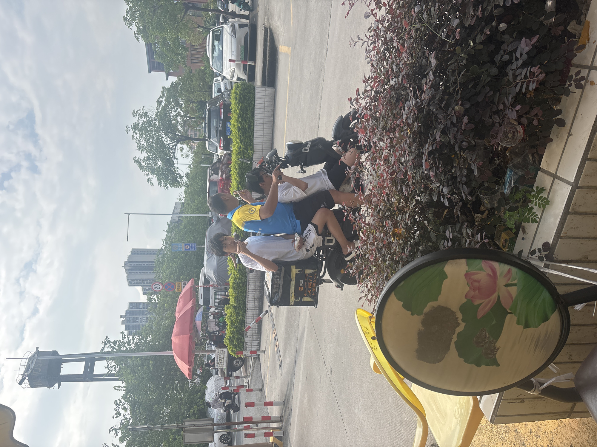 In Nanhai District, China, on a cloudy day, three young males are on an electric scooter on a paved road. Two individuals are seated on the scooter, with the third person appearing to be the driver. The scooter has a large black storage box attached to the rear. The person in the middle is wearing a white t-shirt and shorts, and the person in the back is wearing a blue and yellow t-shirt and black shorts. The front person is wearing a white t-shirt and white shorts. They are all looking in different directions. In the foreground, there is a decorative round fan with a lotus flower painting on it, and to the left, a yellow chair. Behind the scooter, there are cars, trees, buildings, and a red umbrella over a stall. A water tower is visible in the background.