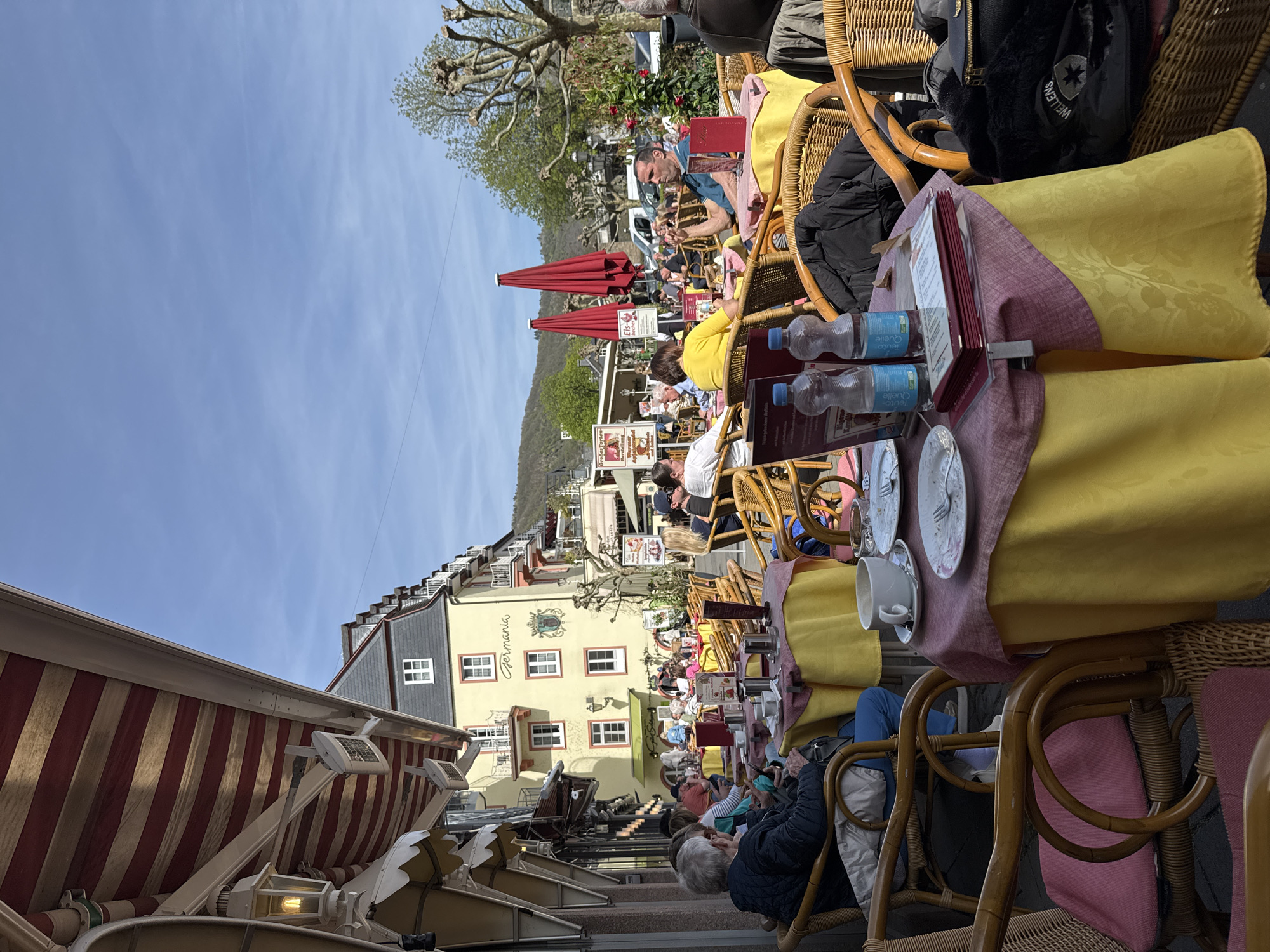 The image shows an outdoor cafe scene in Cochem, Germany. Multiple tables are set with yellow and pink cloths, wicker chairs, plates, cups, water bottles, and menus. People are seated at the tables, eating and socializing. Red umbrellas provide shade. Buildings line the street, including a yellow building with the name "Germania" on it. A striped awning covers the foreground of the cafe seating area. The sky is blue. A small hill is in the background.