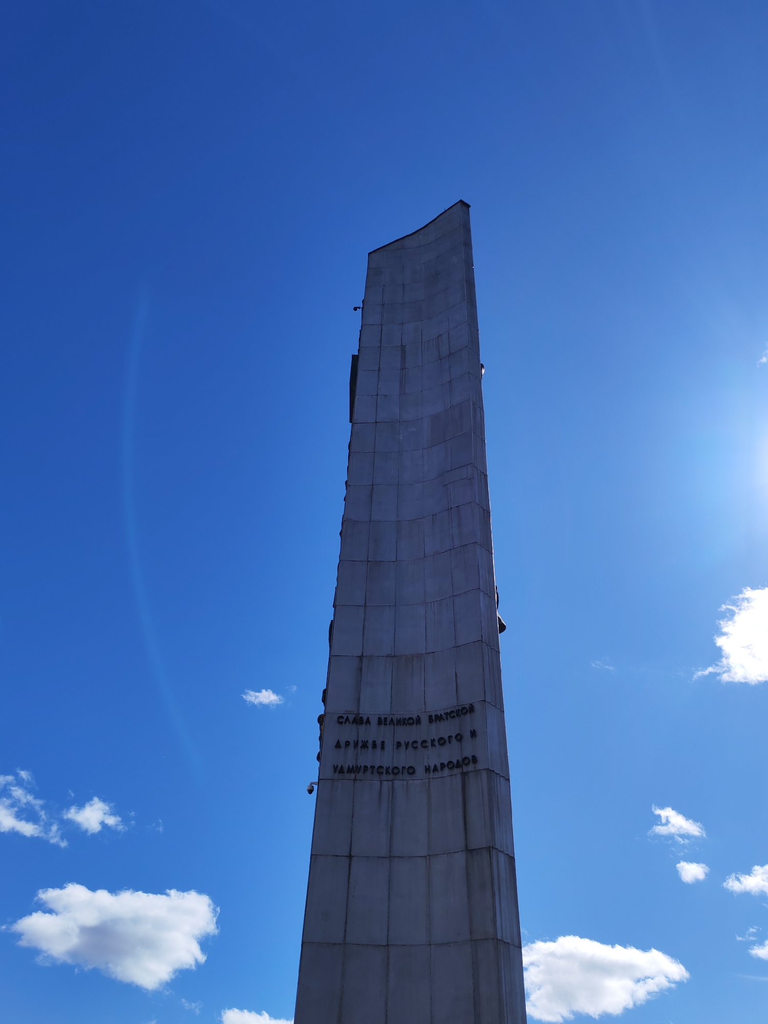 The image shows a tall, gray, monolithic monument under a bright blue sky with scattered white clouds. The monument appears to be made of segmented blocks. Inscribed on its surface are Cyrillic characters arranged in lines. The text is partially obscured by shadow and distance. The sun is visible in the upper right corner, causing a bright glare. The location is Izhevsk, Russia.