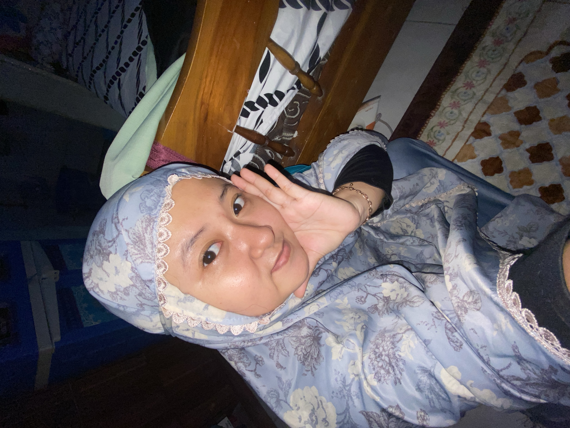 A young Indonesian woman is pictured from the chest up, wearing a light blue hijab with a floral pattern and a lace trim. She is indoors, in what appears to be a bedroom in Gelam, Indonesia. The woman is resting her right hand on her cheek, with her left hand raised slightly in front of her face. She is looking directly at the camera with a gentle smile. To her right, a wooden bed frame is visible, with a patterned blanket and some wooden clips on it. The background is dimly lit, with a hint of a blue cabinet on the left and a patterned rug on the floor. The overall impression is a candid and personal moment captured in a domestic setting.