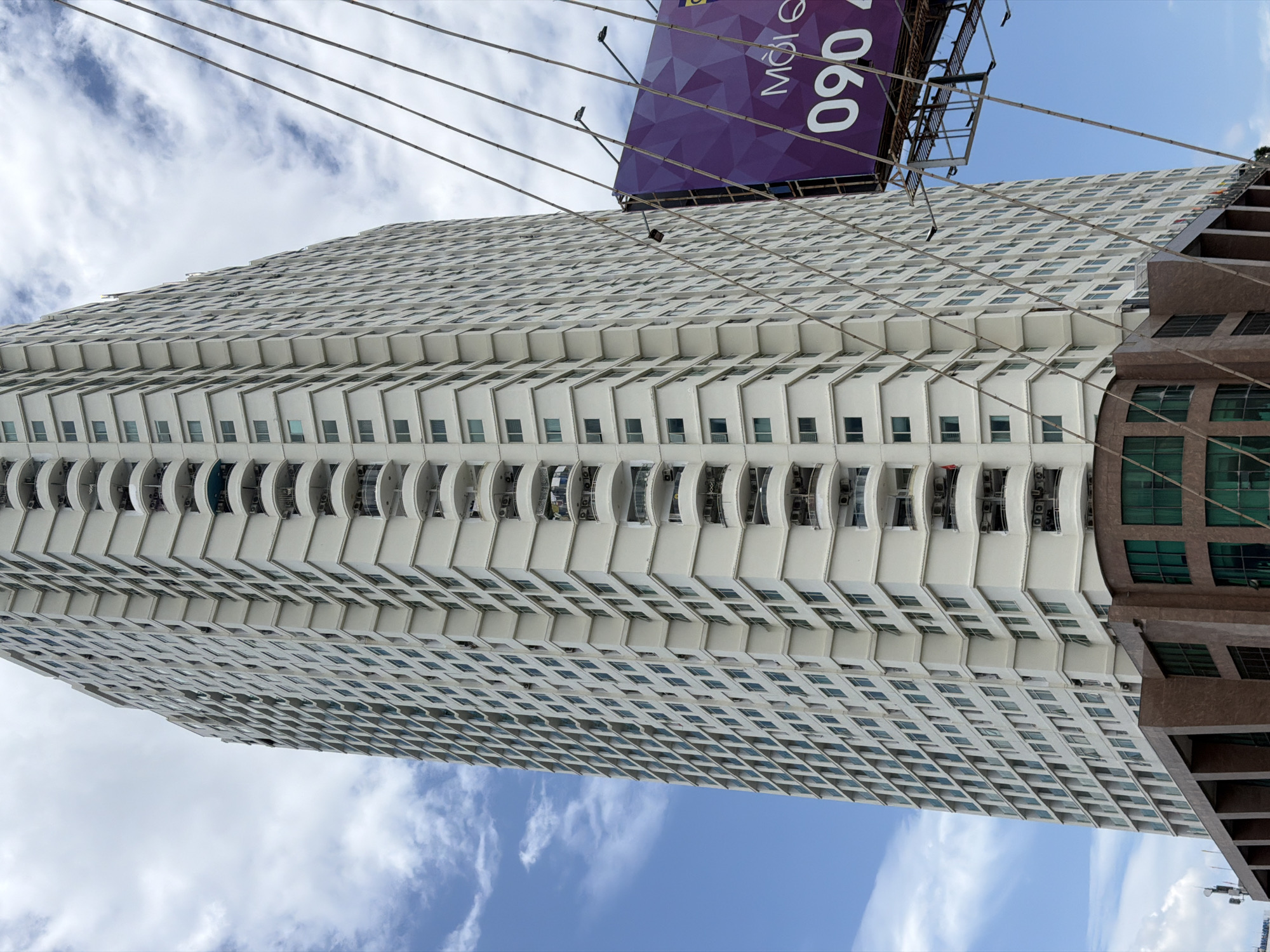 A tall, white apartment building dominates the center of the image, its facade covered in rows of windows and balconies. The building is viewed from a low angle, looking up towards the sky, with a portion of a large purple billboard with white text visible in the right foreground. The billboard appears to be advertising a phone number, "090 4". Several diagonal support wires, possibly from a bridge or construction, are also visible in the foreground, crisscrossing the frame. The sky is a mix of blue with scattered white clouds. The base of the apartment building has a darker, brown facade with green-tinted windows. The scene appears to be in an urban environment, likely Nha Trang, Vietnam. There are no people visible in the image.