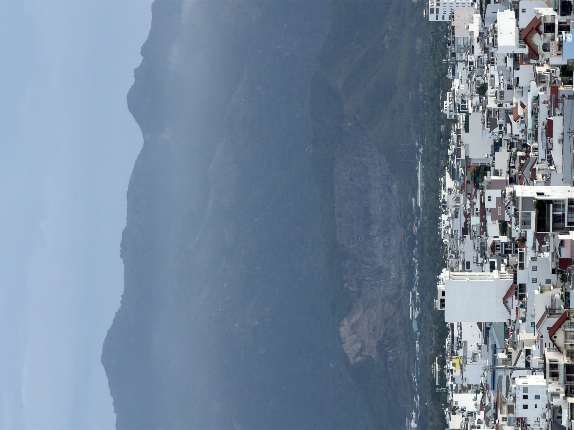 The image displays an elevated panoramic view of a densely built urban area juxtaposed against a large mountain range, consistent with a coastal city like Nha Trang Ward, Vietnam.
In the foreground (right side of the rotated image), a sprawling cityscape features numerous closely packed buildings. These structures are predominantly multi-story and vary in design, with many exhibiting light-colored facades, often white or light grey, contrasted by some terracotta or reddish-brown pitched roofs. The urban density suggests a bustling city environment, with hints of green foliage interspersed among the buildings, likely trees lining streets or within small urban spaces.
Across what appears to be a body of water or a wide expanse of land (not explicitly visible but implied by the separation), the background is dominated by imposing mountains. These mountains are dark, rugged, and appear partially covered in vegetation, showing some rocky outcrops. A hazy atmosphere envelops the mountains, softening their details and contributing to a subdued color palette. The sky above is a light, overcast blue or grey, blending into the distant haze.
The scene depicts a daytime view, although the presence of haze prevents clear, sharp light or distinct shadows, suggesting either an overcast day or atmospheric conditions common in humid coastal regions. There are no people or specific activities discernible within the frame, focusing solely on the natural and built environment. No visible text is present in the image.