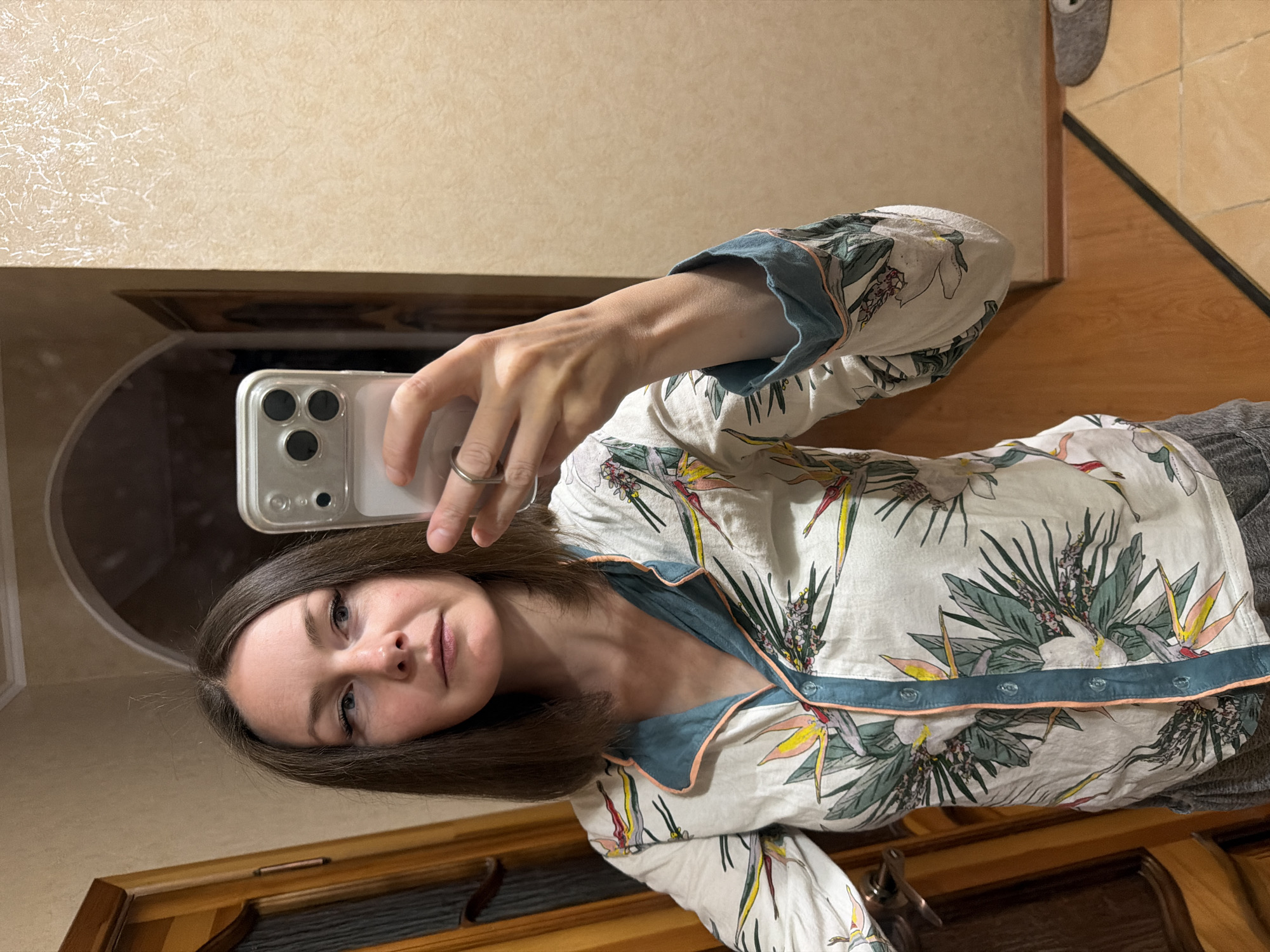 A person is taking a selfie in what appears to be an indoor setting. The individual is wearing pajamas with a floral pattern, holding a smartphone with a clear case in their right hand to capture the image. The background includes a wall with a textured surface and a wooden door frame. The location is unknown.