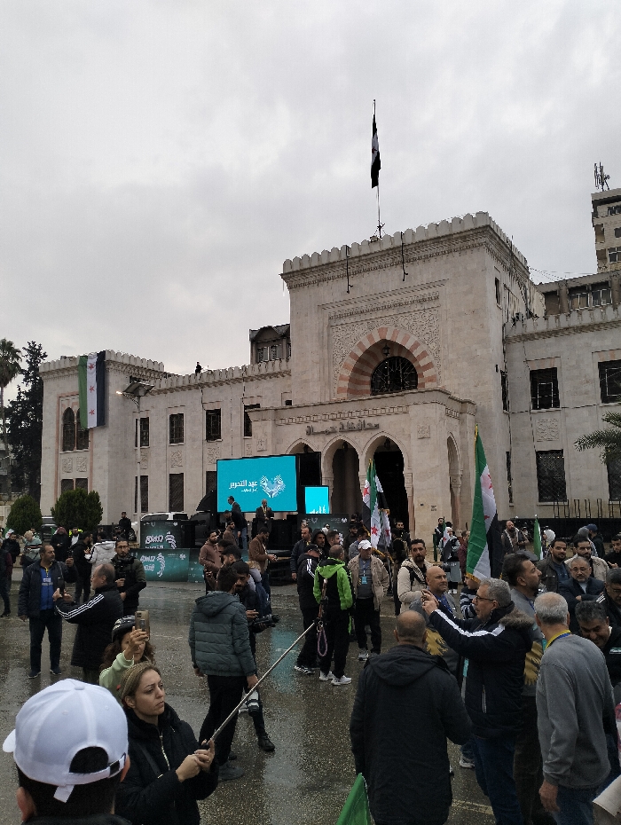 A crowd gathers in front of a large, ornate building beneath a cloudy sky. Several people are holding Syrian flags. A tall screen displaying text in Arabic stands between the crowd and the building's entrance. The architecture of the building includes arches and decorative stonework, topped with a flagpole flying a black and white flag. The ground appears wet, suggesting recent rain.
