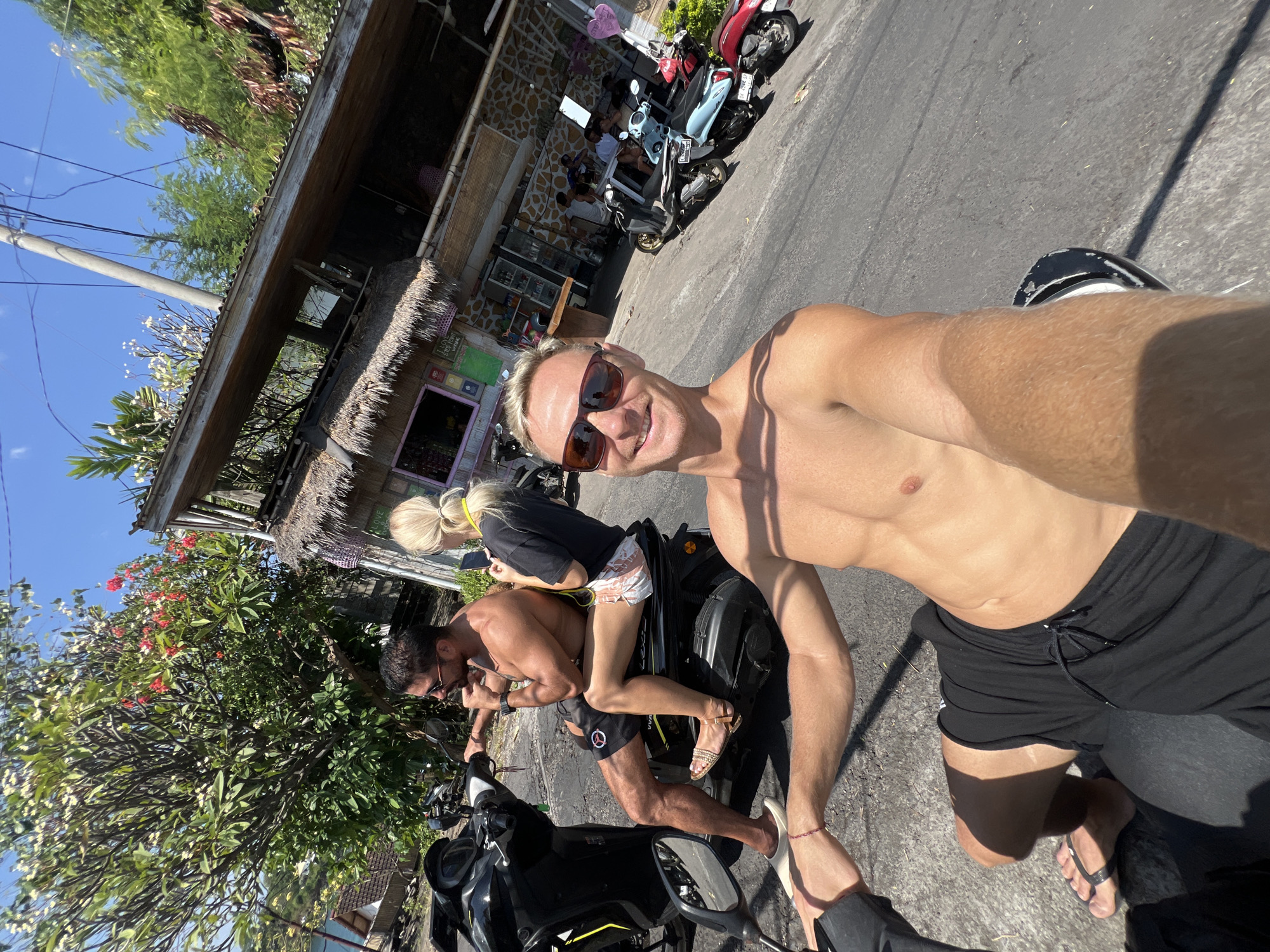 In Abang, Indonesia, a man with blond hair and sunglasses is taking a selfie while riding a scooter. He is shirtless and wearing black shorts. Beside him, a woman with blonde hair pulled back in a ponytail, wearing a black t-shirt and patterned shorts, is seated on the same scooter. Behind them, a man with dark hair and sunglasses, also shirtless and wearing black shorts, is riding another scooter. In the background, there are several buildings with thatched roofs, a tree with red and white flowers, and parked scooters. The scene appears to be on a sunny day, with a clear blue sky and overhead wires visible.