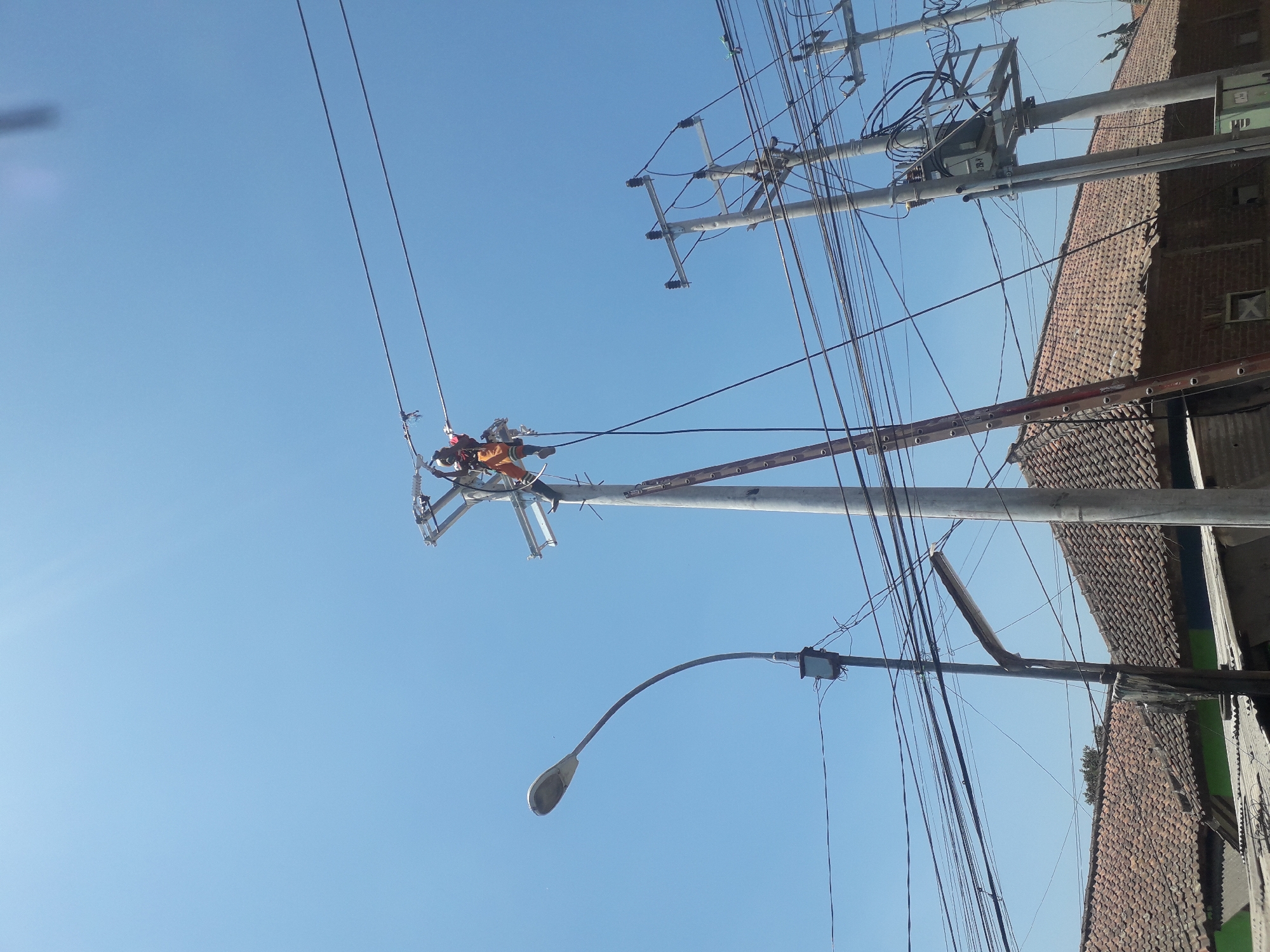 In Cankuang, Indonesia, a lineman wearing an orange safety vest and black pants is working on a tall utility pole. He is positioned near the top of the pole, attached to equipment, and appears to be manipulating electrical wires. A red ladder leans against the pole, providing access to his work area. Below the lineman's pole, a street light stands with its curved arm extending to the left. The scene is framed by a clear blue sky and the roofs of several buildings with red clay tiles. In the background, more utility poles with transformers and a dense network of electrical wires are visible, indicating a populated urban or suburban area.
