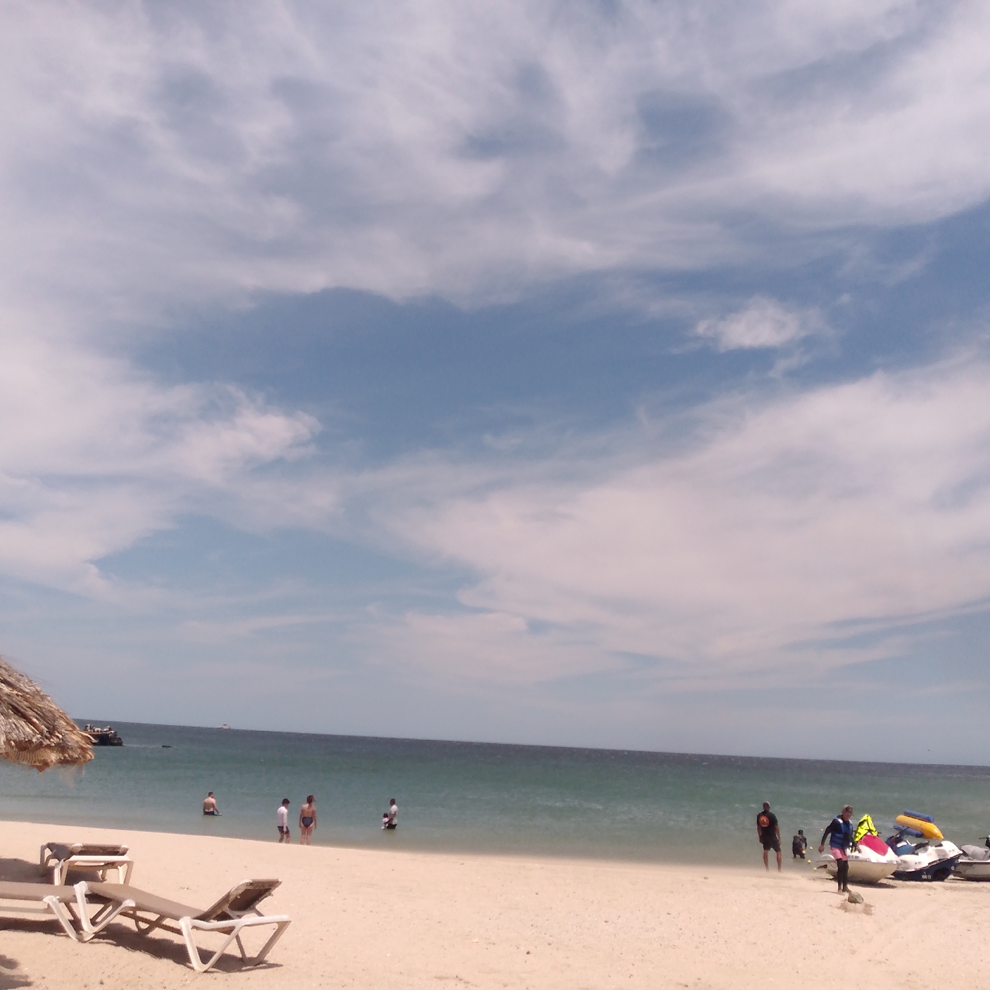 The image displays a vibrant daytime beach scene in Pedro González, Venezuela, characterized by a wide expanse of ocean and sandy shore under a partially cloudy sky. White, diffuse clouds are scattered across the blue sky.
The tranquil, blue-green ocean meets a light-colored sandy beach. In the foreground on the left, several white lounge chairs are arranged on the sand, with one showing a person's legs. A straw-thatched umbrella is visible on the far left, providing a tropical accent. In the mid-ground, several individuals are seen enjoying the shallow waters of the sea; some are wading, and one person appears to be partially submerged. Distant boats or structures are visible on the horizon line.
On the right side of the beach, a group of people is gathered around two or more jet skis resting on the sand, some equipped with colorful yellow and blue inflatable tubes. One person wears a life vest, indicating preparation for or participation in water sports. Other individuals are standing near the jet skis or in the adjacent shallow water.
The scene captures a leisurely and recreational atmosphere typical of a tropical beach destination. The time of day appears to be mid-day or early afternoon due to the bright, though diffused, lighting. No legible text is visible on any objects or clothing in the image.