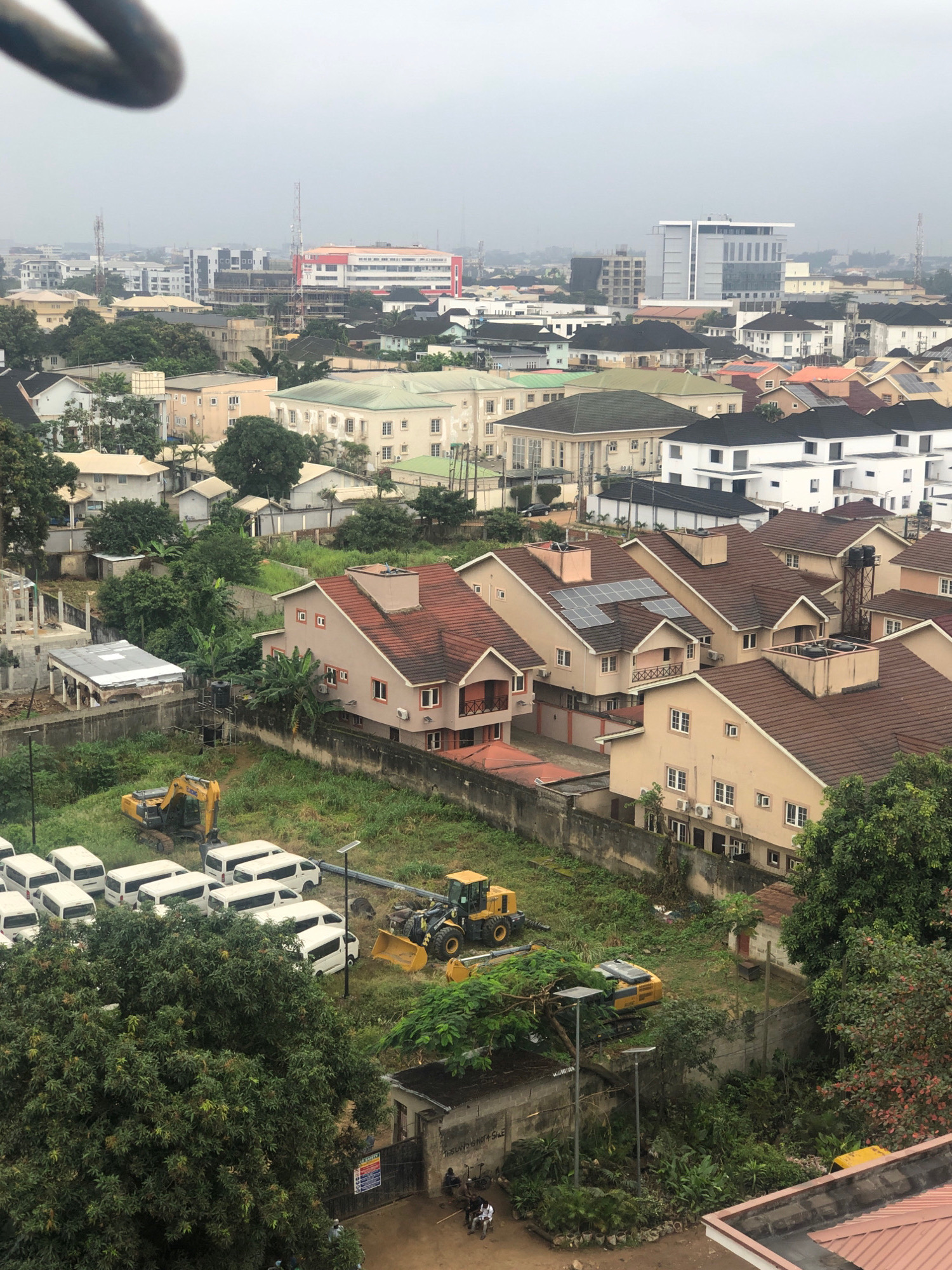 This is a high-angle, medium shot photograph of a residential and commercial area in Ikeja, Nigeria, taken on a cloudy day. In the foreground, the top of a curved, dark object, possibly a railing or pipe, is visible. The midground reveals a cluster of two-story and three-story buildings with red-tiled roofs and cream-colored exteriors. Some of these buildings have solar panels installed on their roofs. A few of the buildings have visible air conditioning units mounted on their walls. In the lower left quadrant of the image, a line of white vans is parked on a grassy area. A yellow excavator is positioned in the grass between the vans and a red-tiled building. Another yellow construction vehicle, a front-end loader, is also present in the grassy area. The background shows a cityscape with taller buildings, including a modern, gray skyscraper and a red and white building with a prominent antenna. There are also numerous other residential buildings and trees scattered throughout the scene. The overall impression is one of urban development with ongoing construction or maintenance activities.